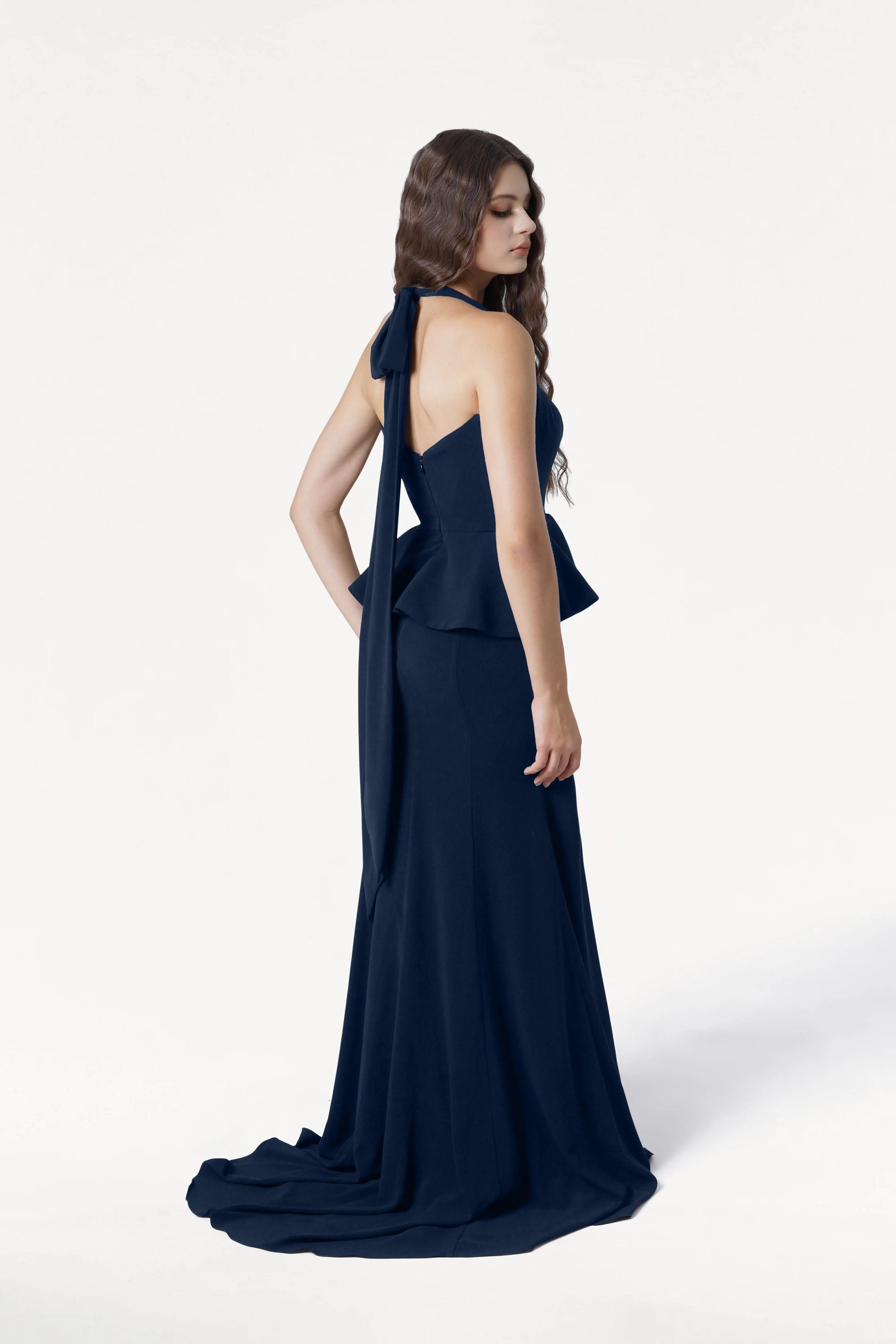 A woman with long wavy hair stands in profile wearing the TH&TH Peony Bridesmaid Dress in Navy, a modern floor-length gown featuring moss crepe fabric, a peplum waist, and bow-tied back strap, against a plain white background.