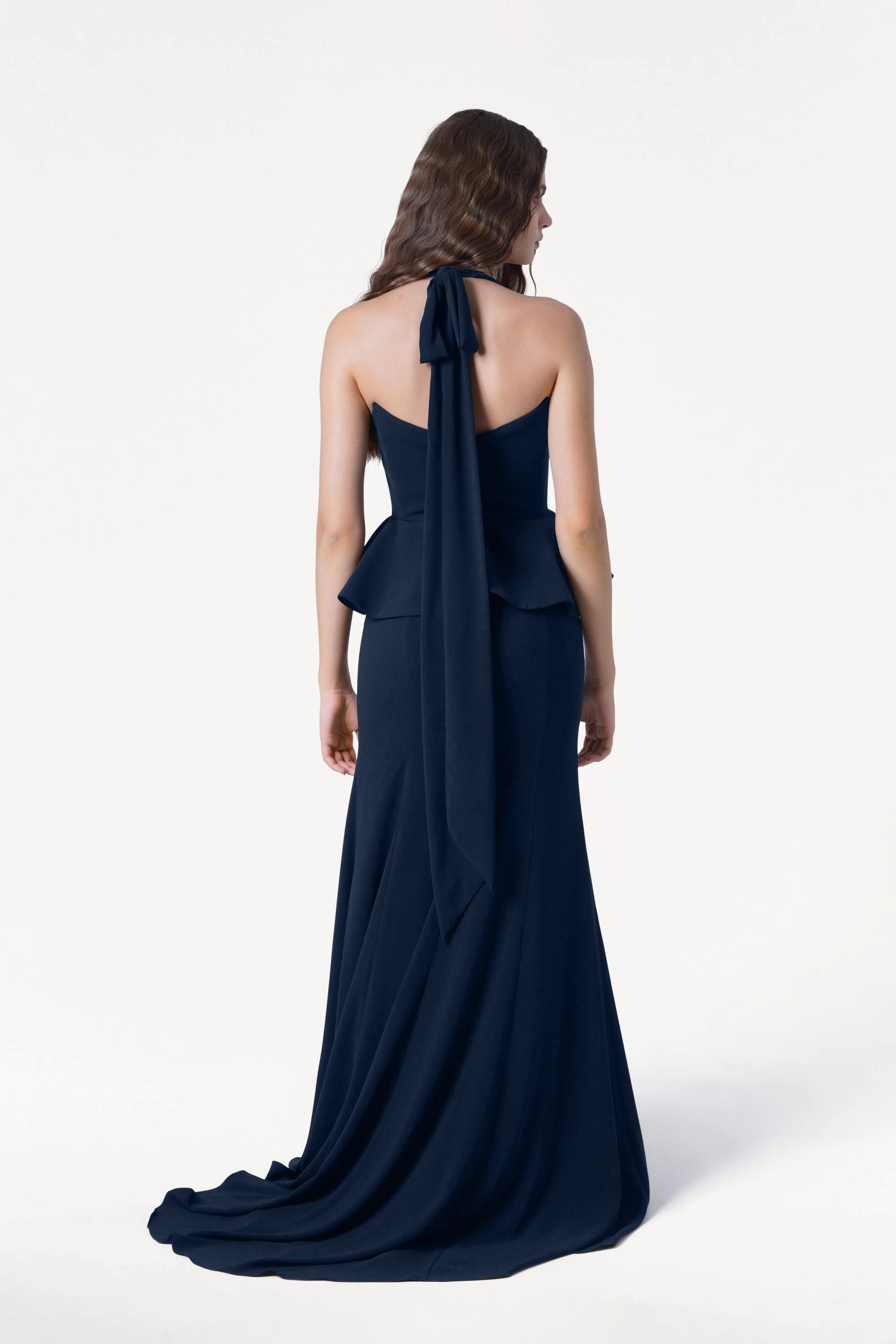 A woman with long wavy hair faces away, wearing the TH&TH Peony Bridesmaid Dress in Navy—a floor-length, size-inclusive gown with a peplum waist and long sash—set against a plain white background.