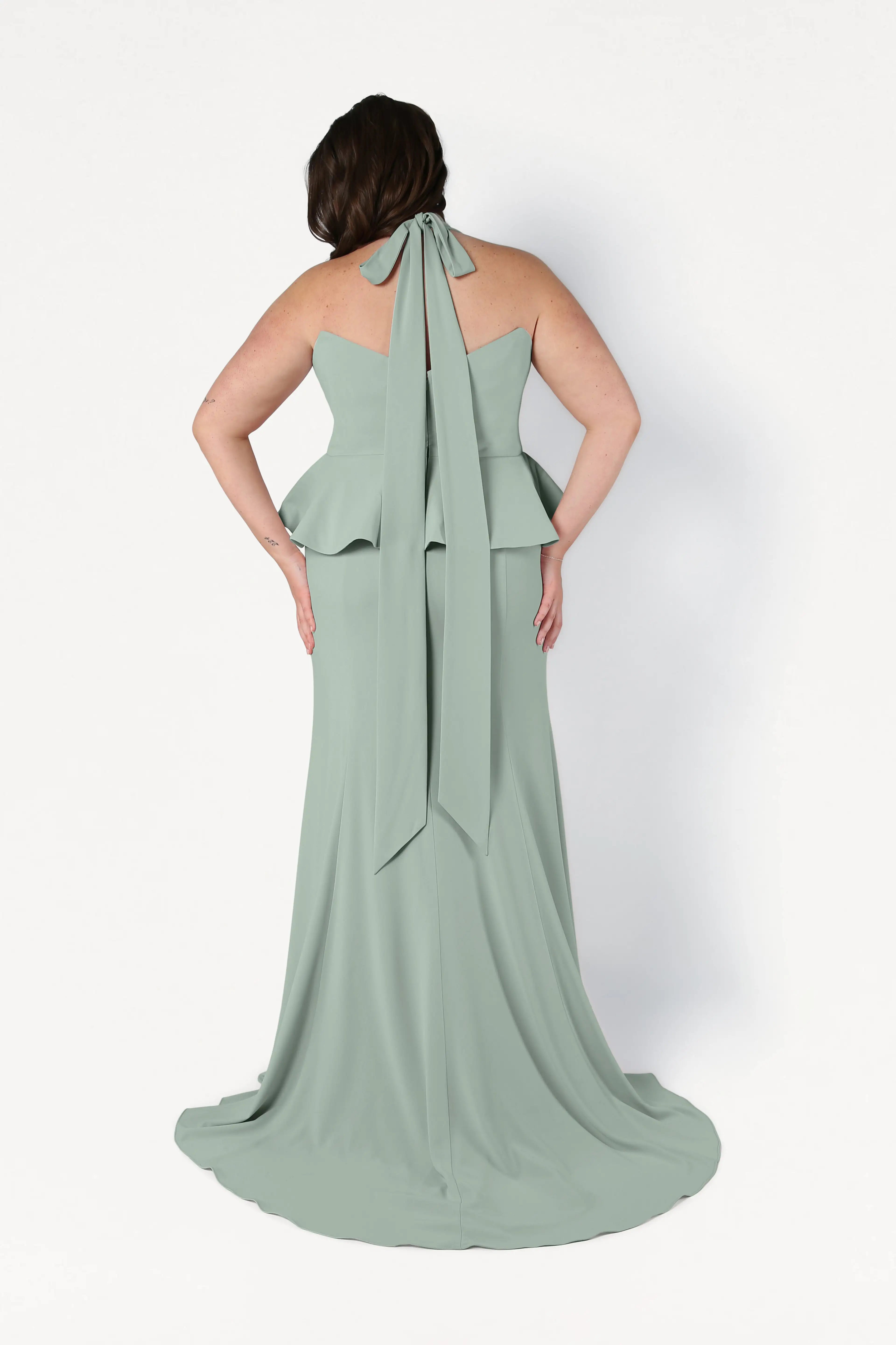 A woman faces away, showcasing the back details of the TH&TH Peony Bridesmaid Dress in Sage Green, a floor-length gown with a peplum design and long neck tie, set against a plain white background.