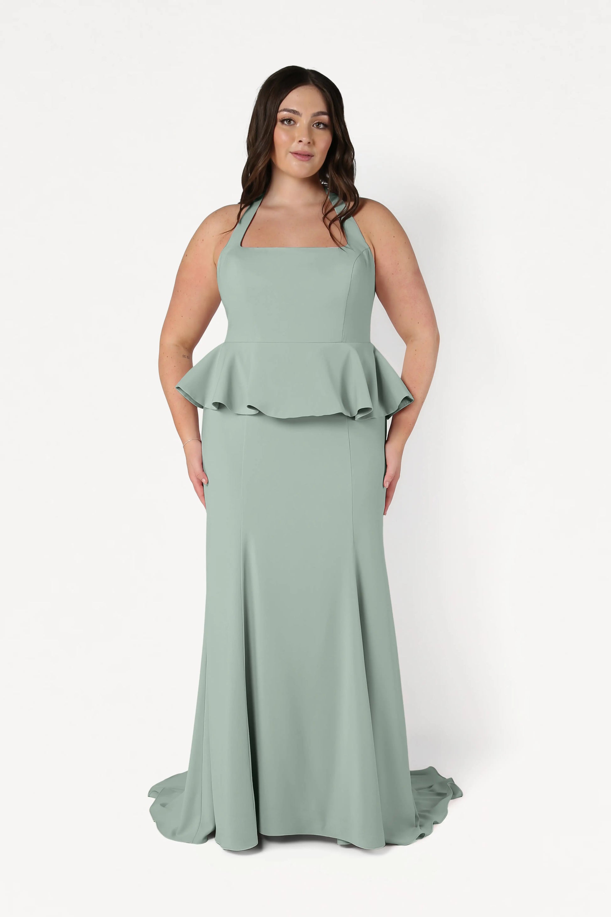 A woman with long dark hair and a neutral expression models the TH&TH Peony Bridesmaid Dress in Sage Green, featuring moss crepe fabric, a square neckline, halter straps, and a peplum waist, against a white background.