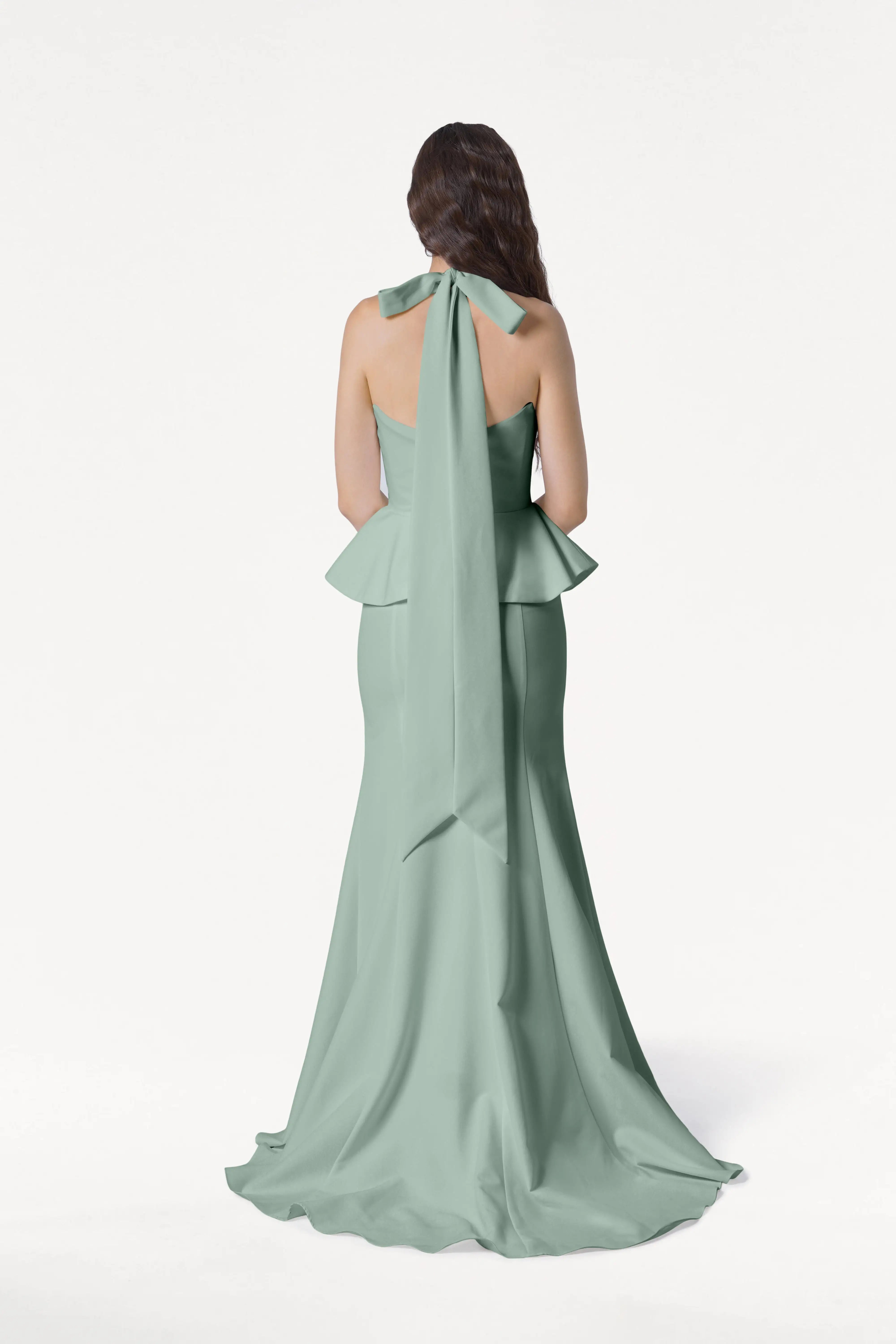 A woman with long, wavy brown hair models the TH&TH Peony Bridesmaid Dress in Sage Green—a floor-length peplum gown with a neck bow—standing against a plain white background, facing away from the camera.
