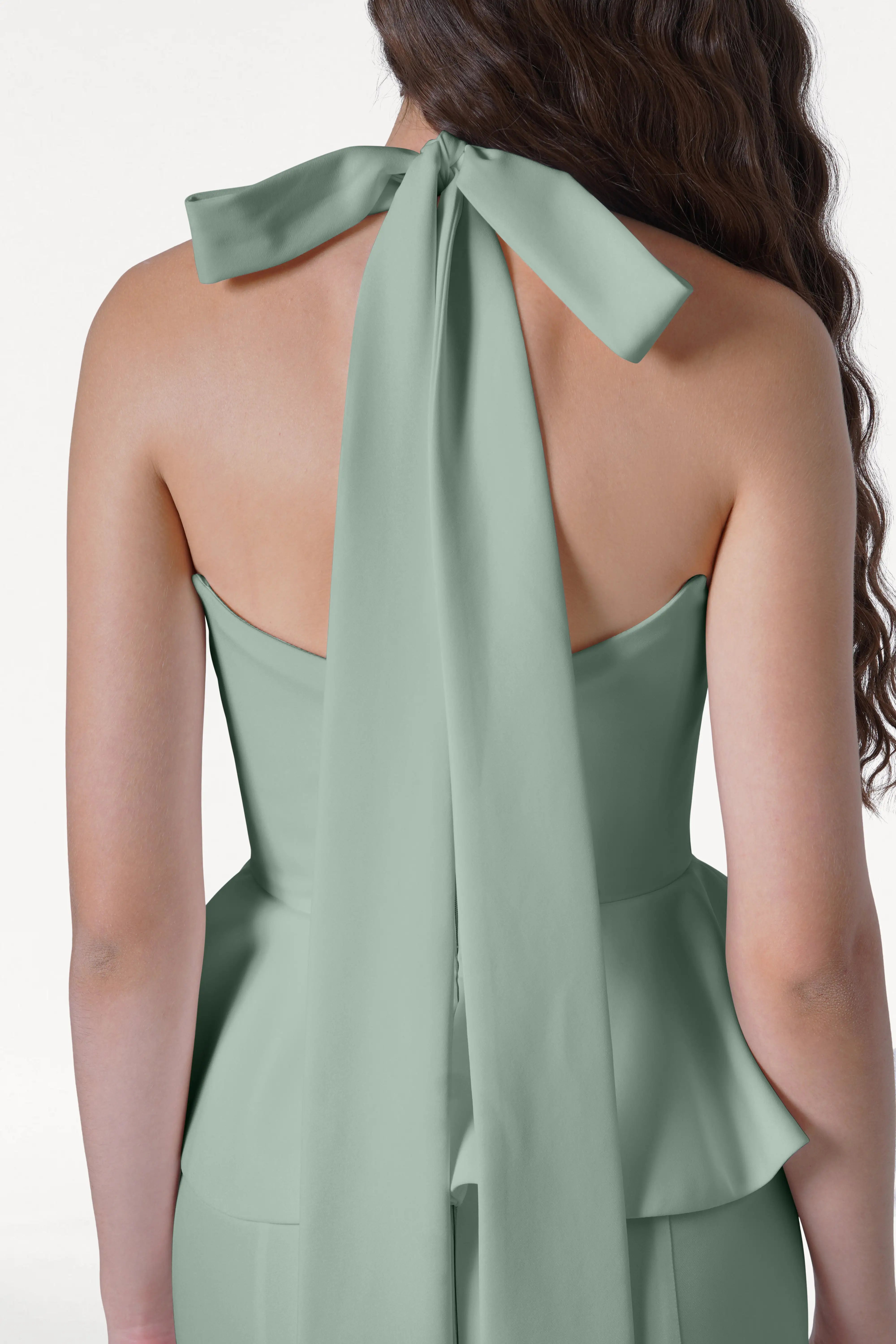 A woman with long wavy brown hair is seen from the back wearing the TH&TH Peony Bridesmaid Dress in Sage Green—a modern moss crepe halter dress featuring a large bow, long ribbon ties, a fitted silhouette, and a peplum hem.