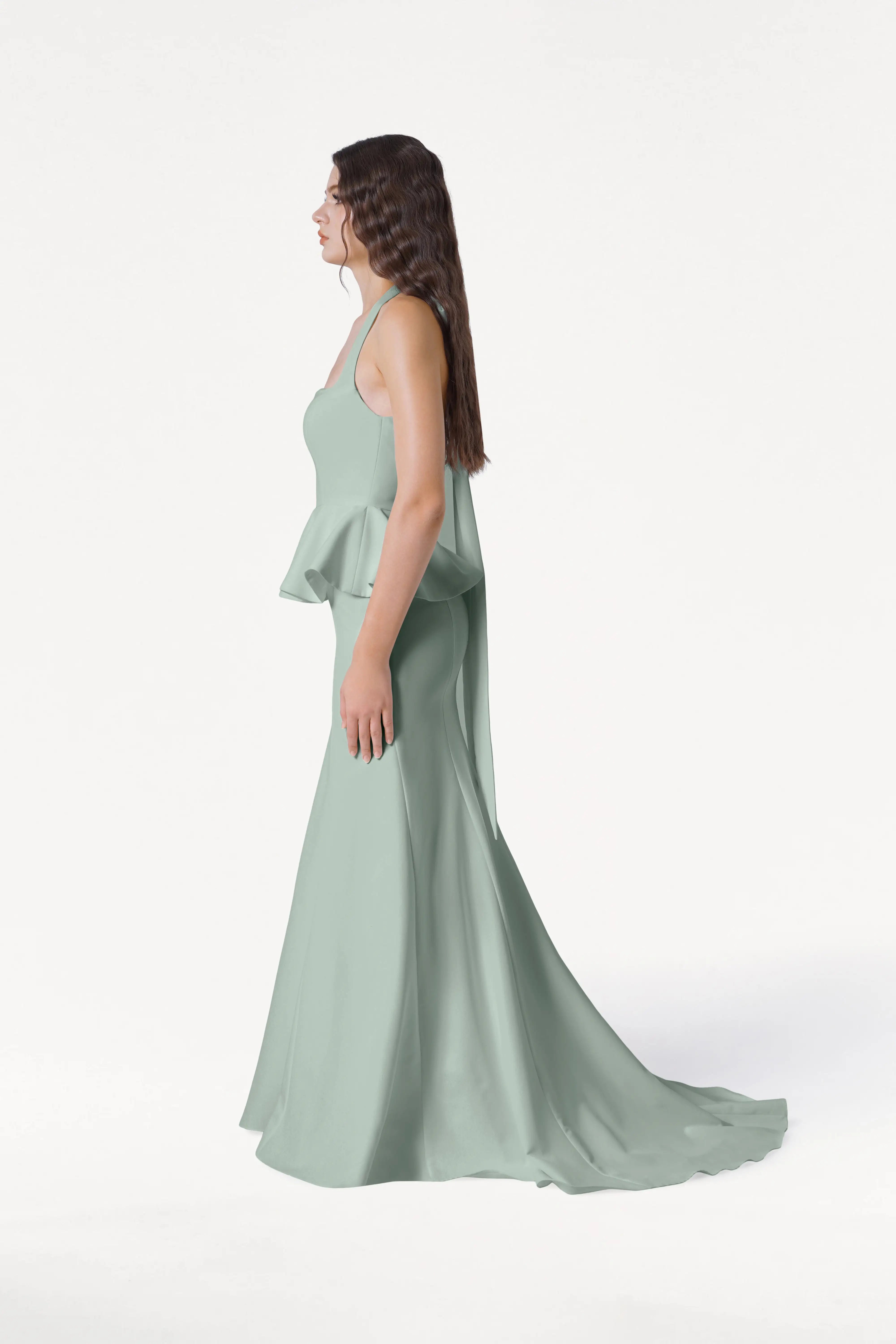 A woman stands in profile against a white background wearing the TH&TH Peony Bridesmaid Dress in Sage Green, a modern crepe gown with a fitted bodice and flowing skirt with train for an elegant look.