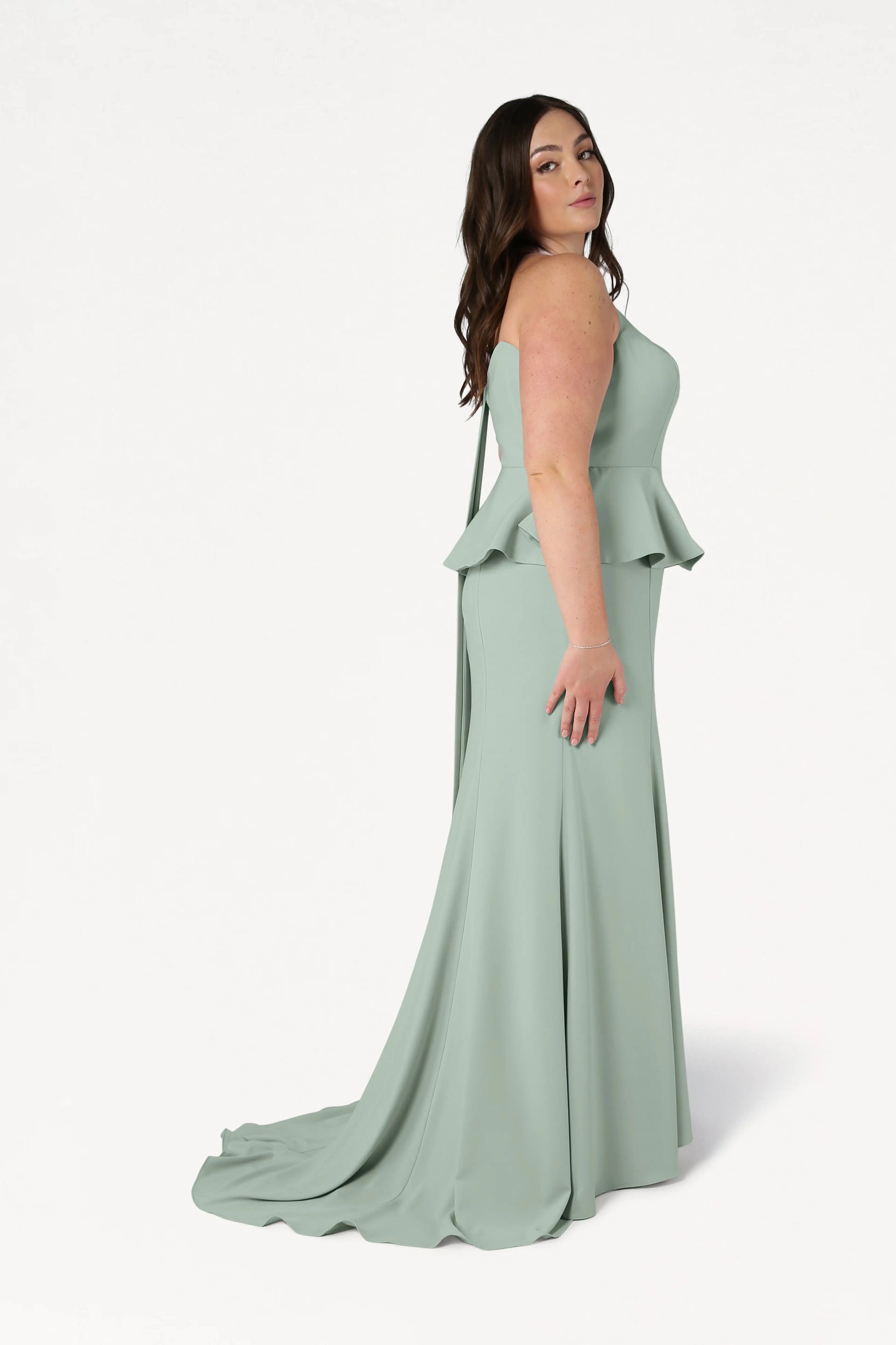 A woman with long brown hair stands sideways against a plain white background, wearing the TH&TH Peony Bridesmaid Dress in Sage Green—a modern strapless moss crepe gown with a peplum and floor-length skirt.