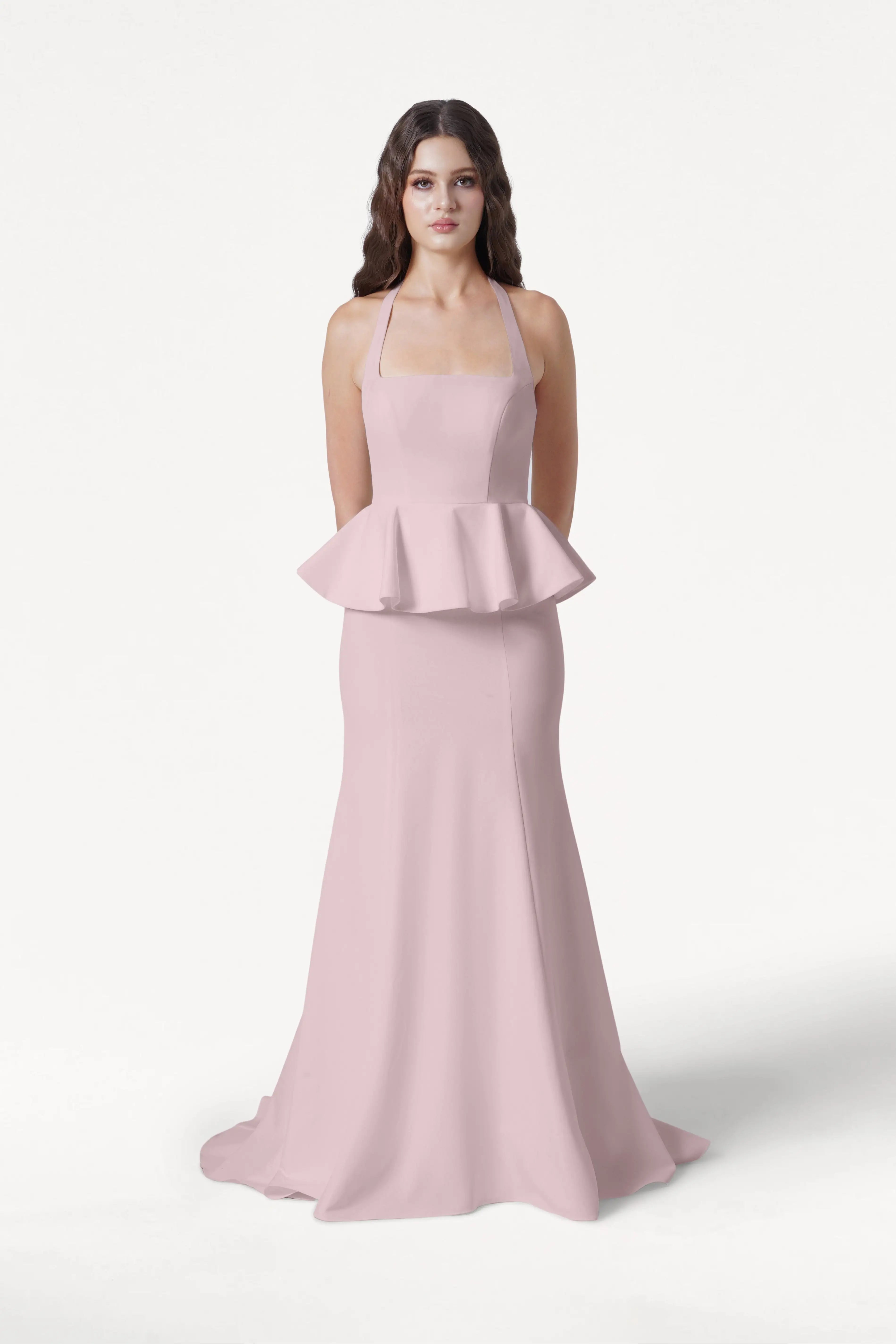 A woman with long brown hair stands against a white background, wearing the TH&TH Peony Bridesmaid Dress in Smoked Blush—a sleeveless gown with a peplum waist and floor-length skirt. Her hands are behind her back.