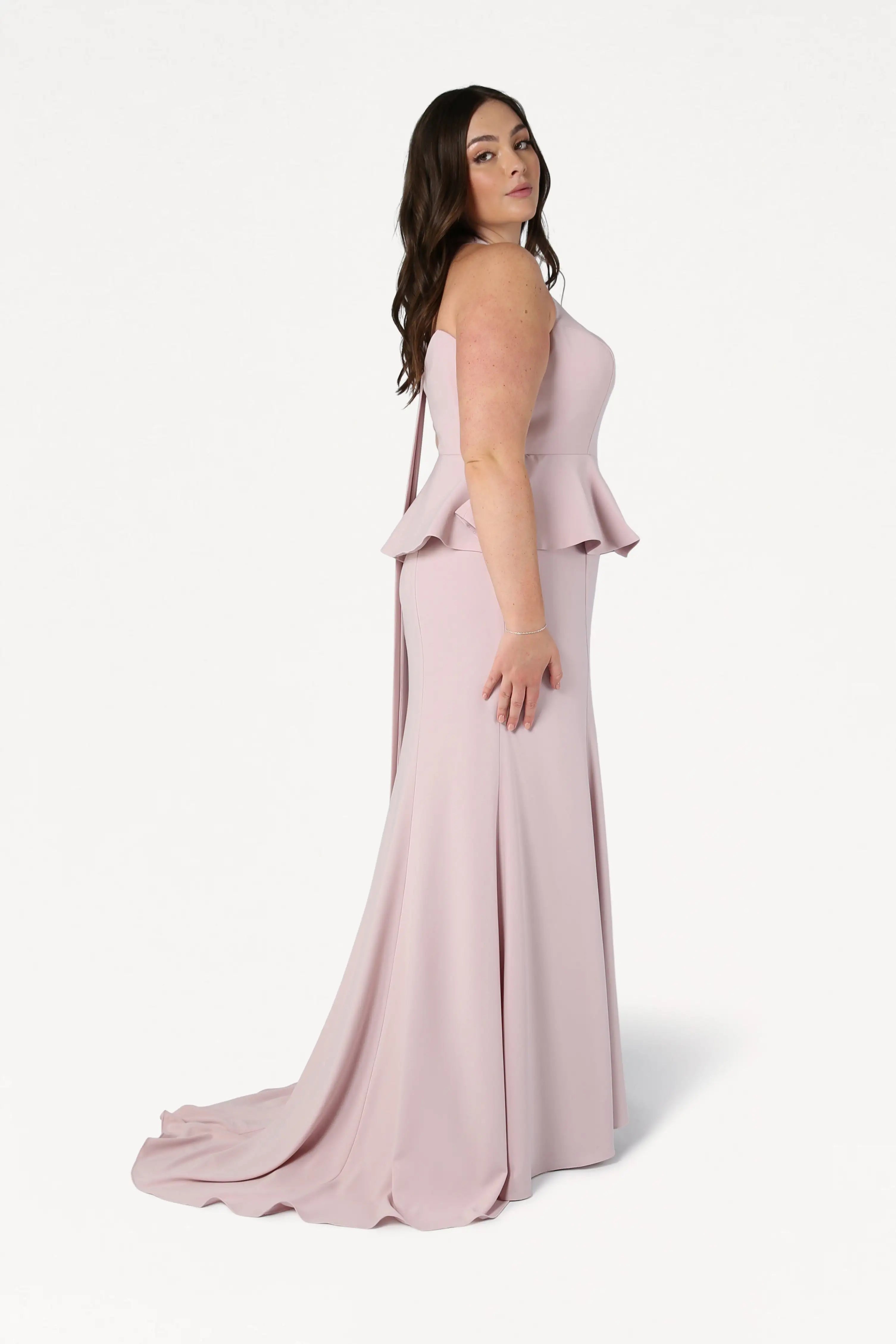 A woman with long brown hair models the TH&TH Peony Bridesmaid Dress in Smoked Blush, a size-inclusive, strapless gown with a fitted bodice and floor-length flared skirt, against a plain white background.