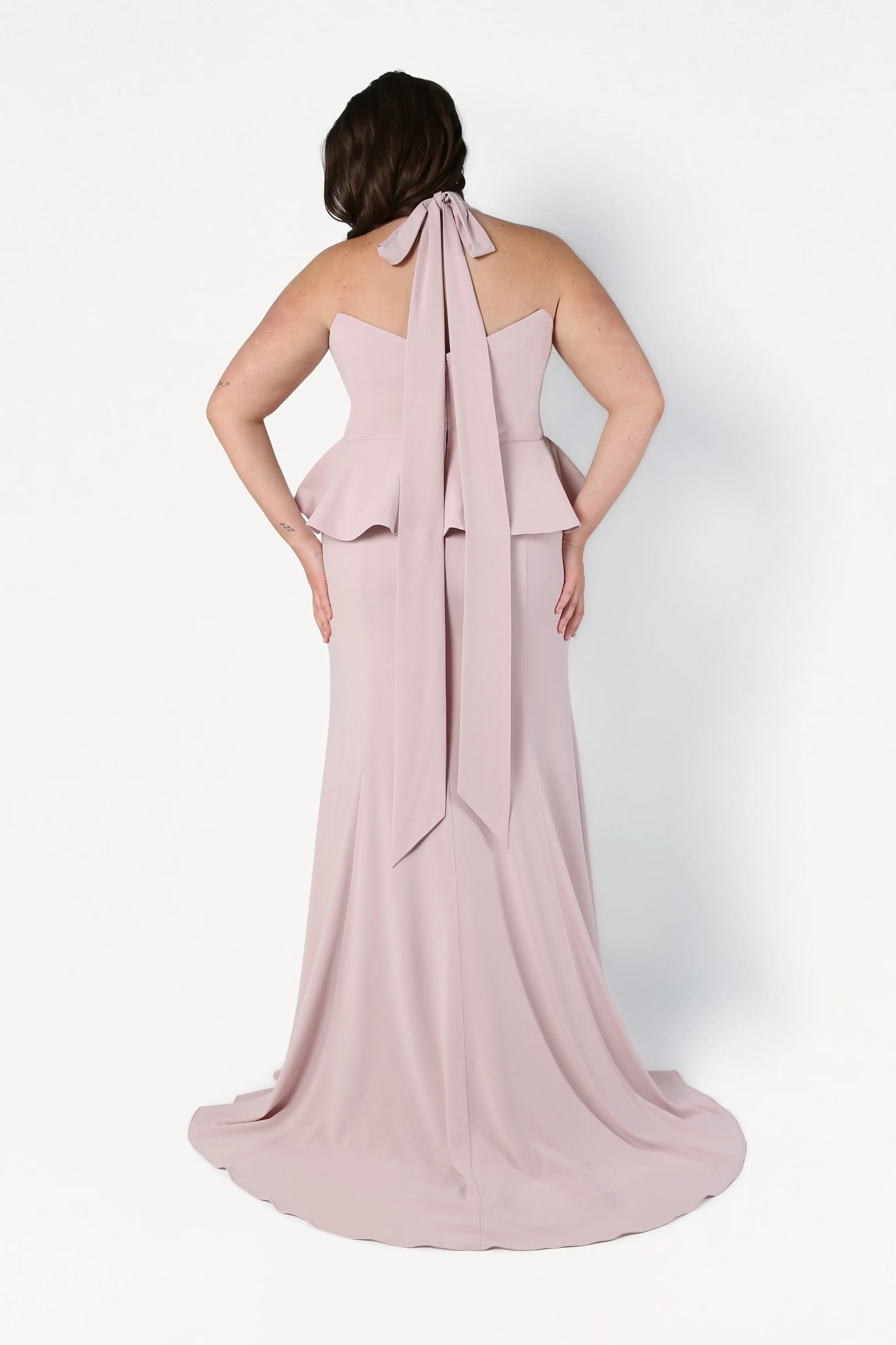 Woman wearing a pink halter neck gown with a large bow detail on a white background