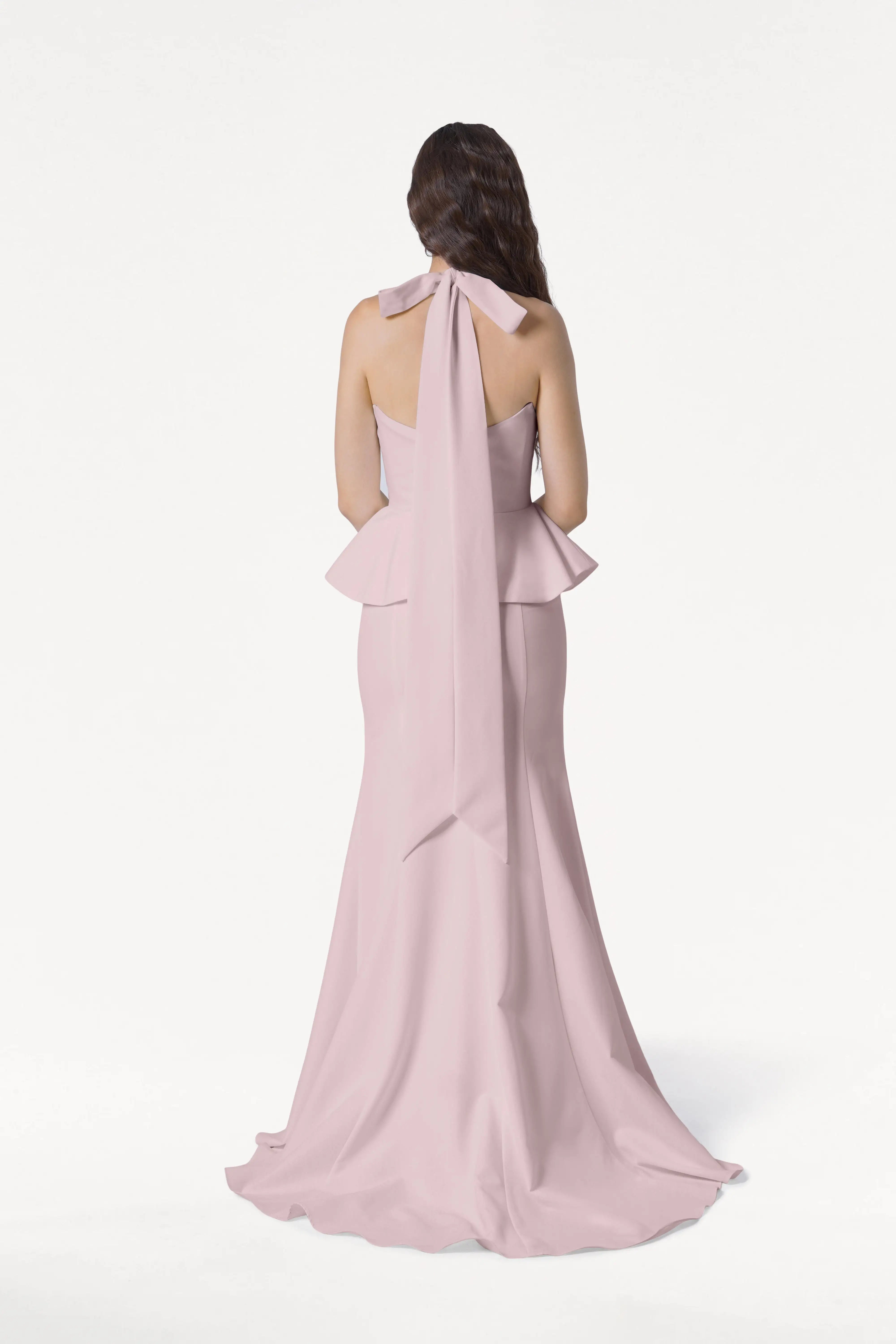 A woman with long brown hair stands facing away, wearing the TH&TH Peony Bridesmaid Dress in Smoked Blush—a modern strapless gown with a ruffled peplum waist and a long bow at the back. The background is plain white.
