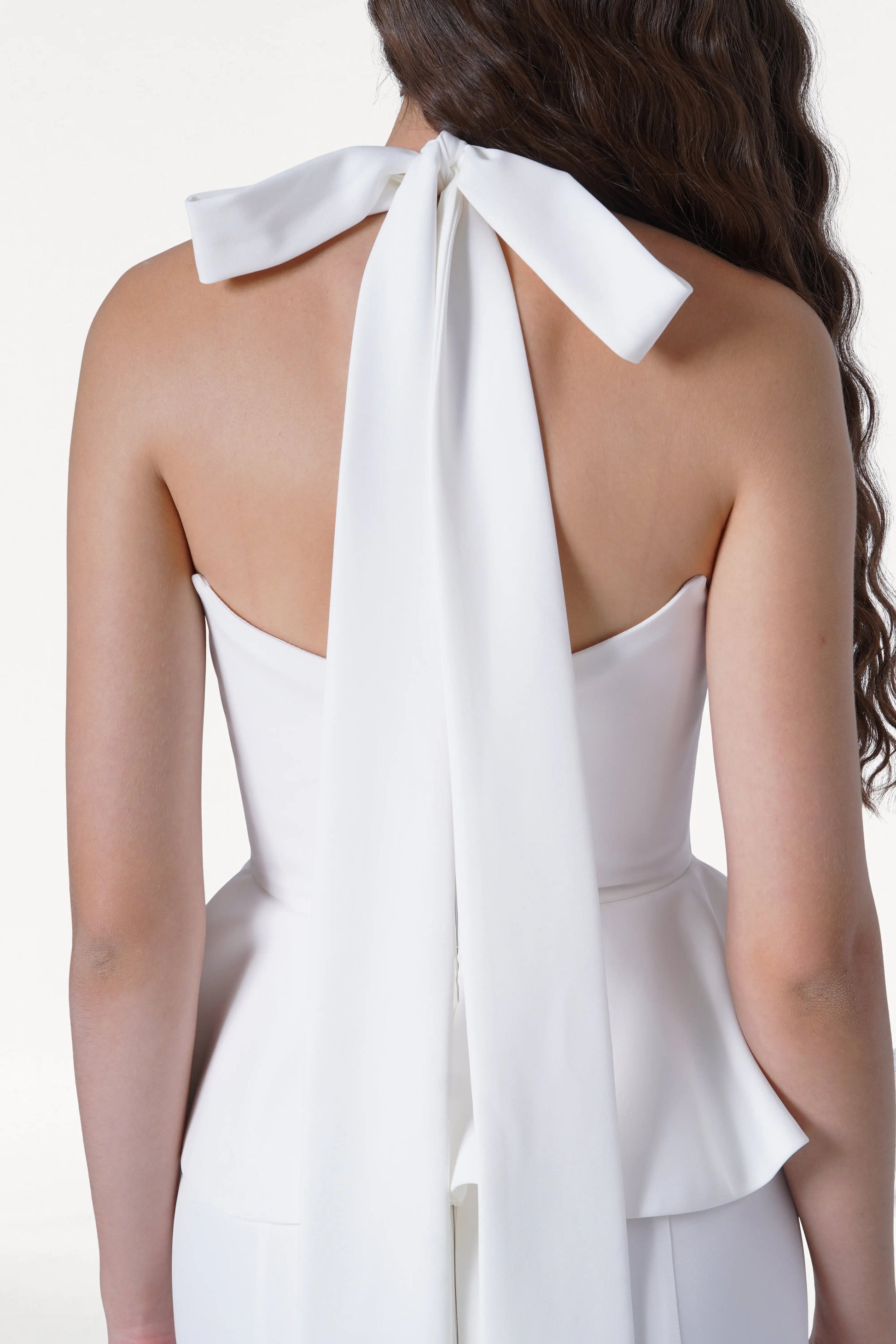 A woman with long, wavy brown hair is shown from the back wearing the TH&TH Peony Dress in Ivory—a modern moss crepe bridesmaid dress featuring a strapless design with a large bow and long neck ribbon.
