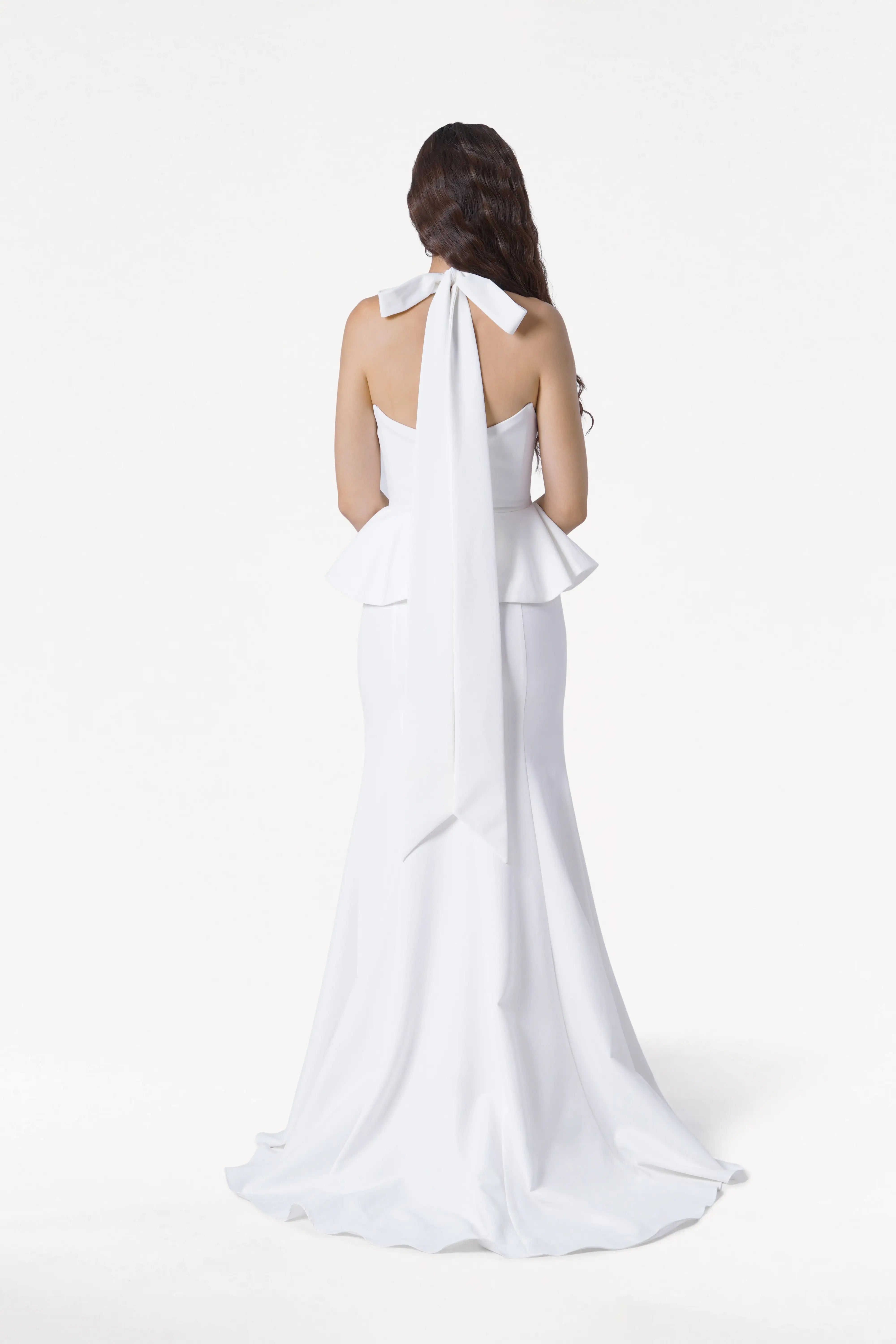A woman with long, wavy brown hair stands facing away in the TH&TH Peony Dress in Ivory—a sleeveless, floor-length moss crepe dress featuring a peplum waist and large bow at the neck. The background is plain white.