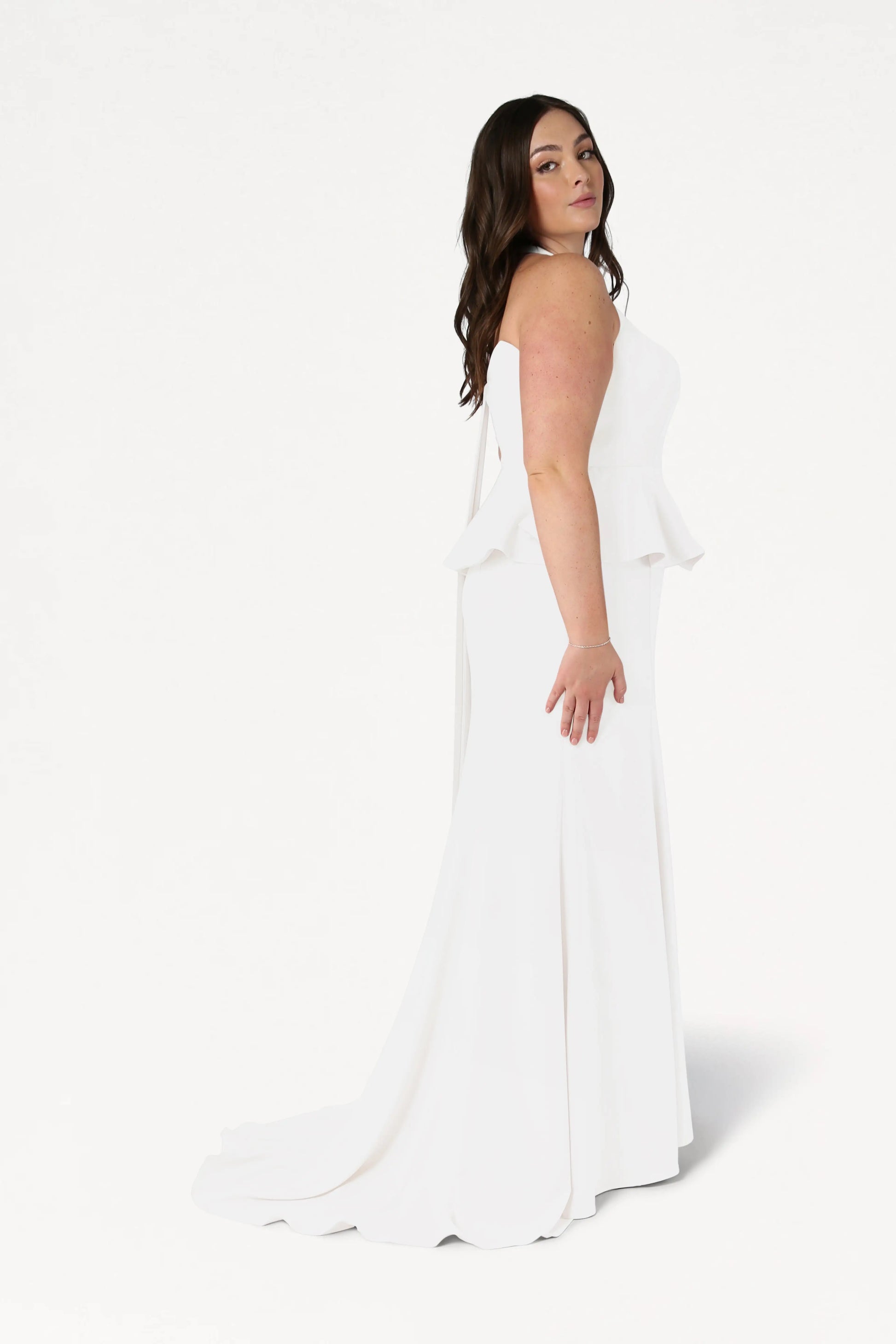 A woman with long brown hair stands in profile against a plain white background, wearing the TH&TH Peony Dress in Ivory—a size-inclusive, strapless, fitted gown with peplum detail.
