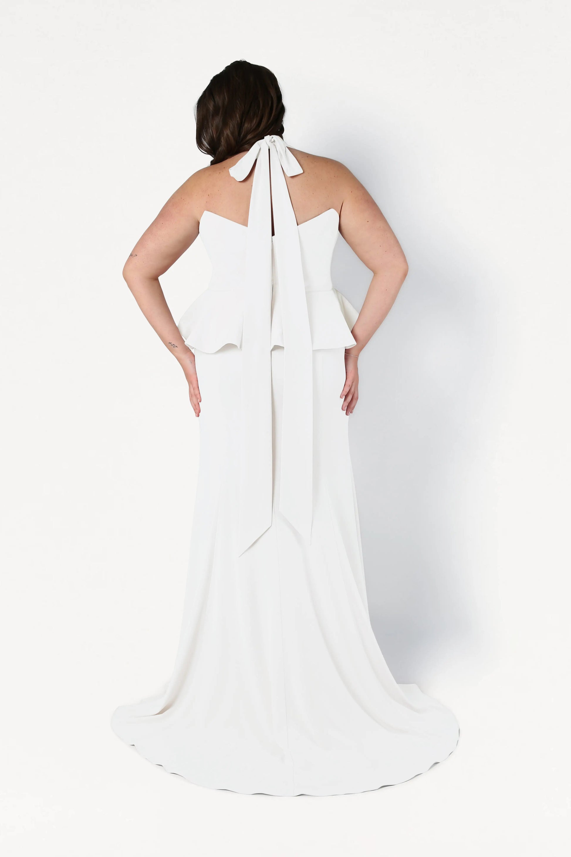 A woman with brown hair, facing away, wears the TH&TH Peony Dress in Ivory—an inclusive-size, floor-length halter gown with a large back bow and waist peplum. The background is plain white.