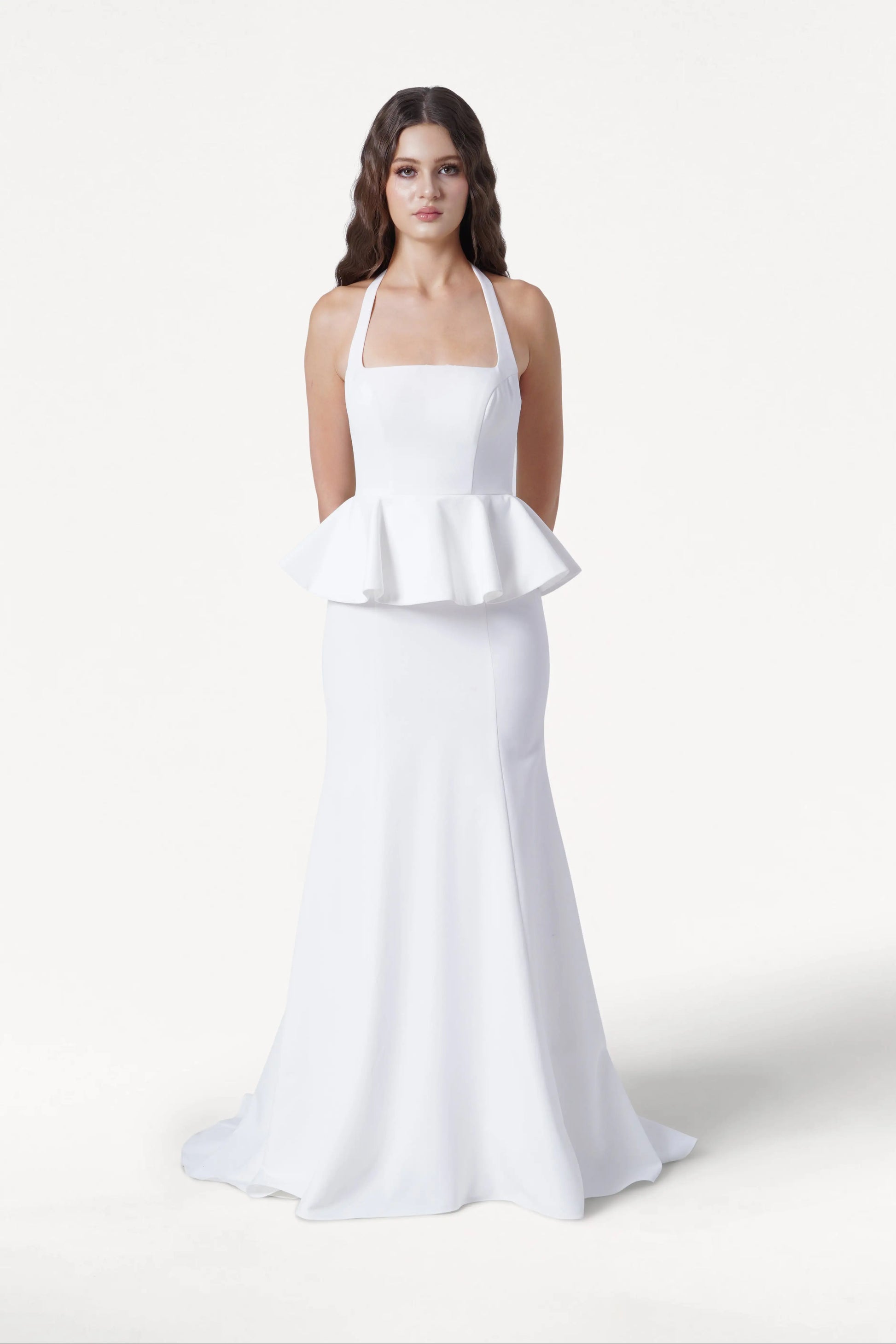 A woman faces forward in the TH&TH Peony Dress in Ivory, a size-inclusive modern moss crepe bridesmaid gown featuring a long, white sleeveless silhouette, halter neckline, and waist peplum detail. The background is plain white.
