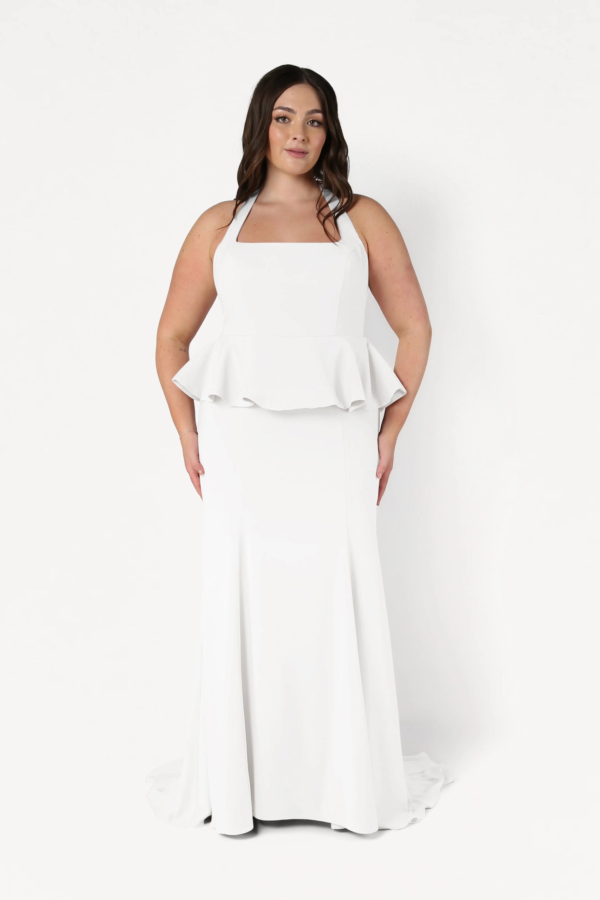 A woman with long dark hair models the size-inclusive Peony Dress in Ivory by TH&TH, featuring a white halter-neck, peplum waist, and floor-length skirt, standing against a plain white background.