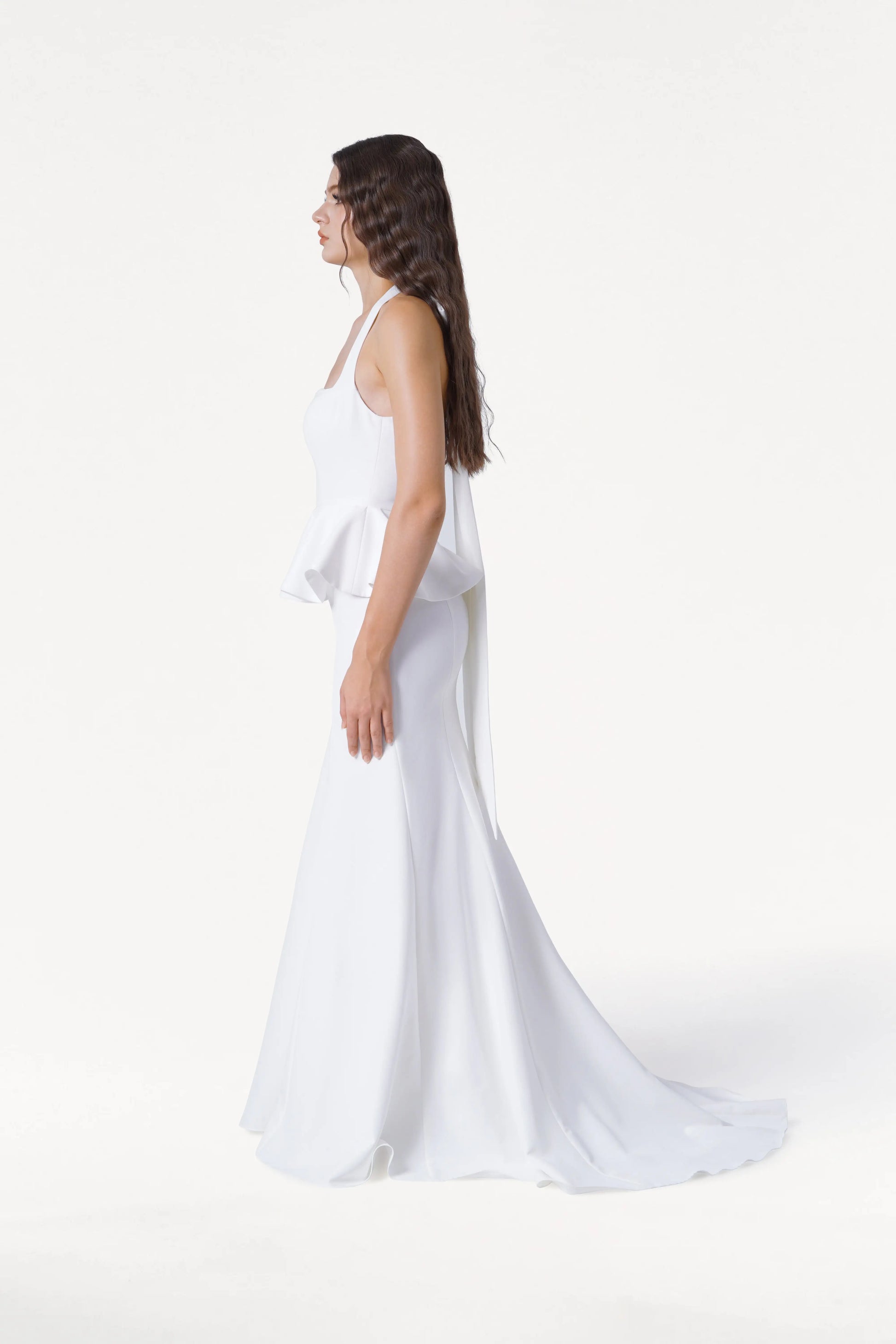 A woman in profile stands against a white background, wearing the TH&TH Peony Dress in Ivory—a sleeveless, floor-length moss crepe gown with peplum detail. Her long, wavy hair cascades down her back.