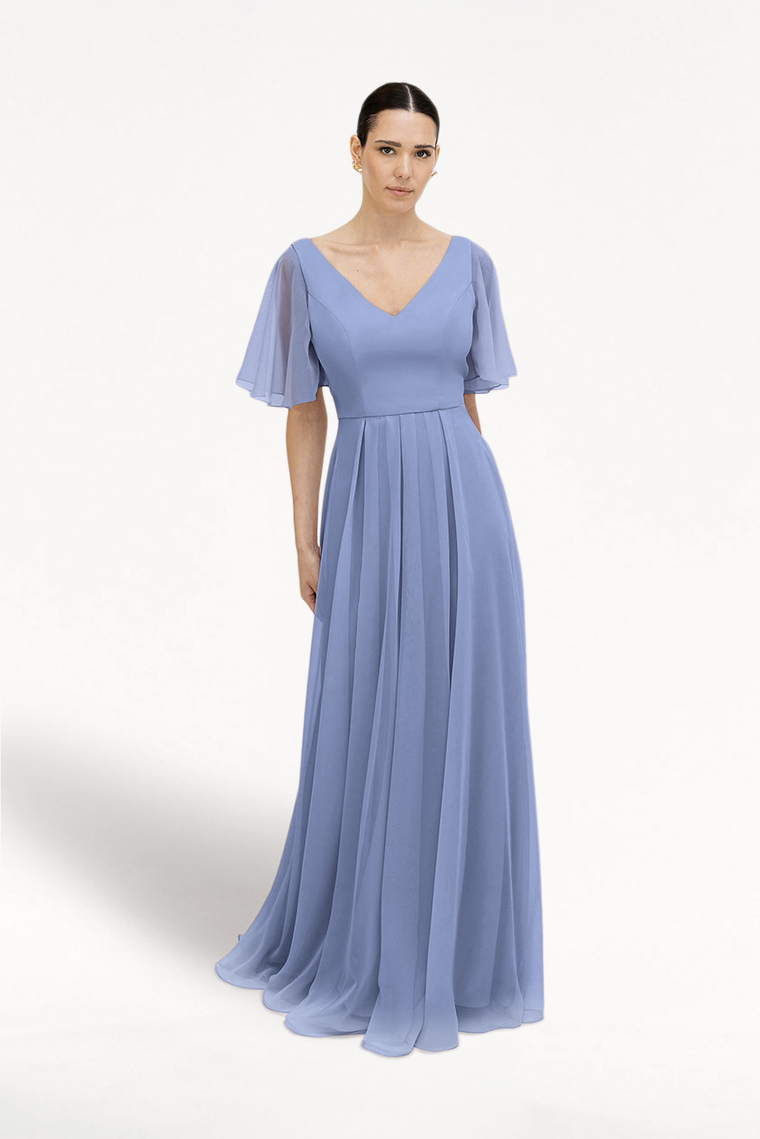 A woman wears the TH&TH Phoebe Bridesmaid Dress in French Blue chiffon, featuring short flutter sleeves, V-neckline, fitted waist, and a flowing pleated floor-length skirt, standing against a plain white background.