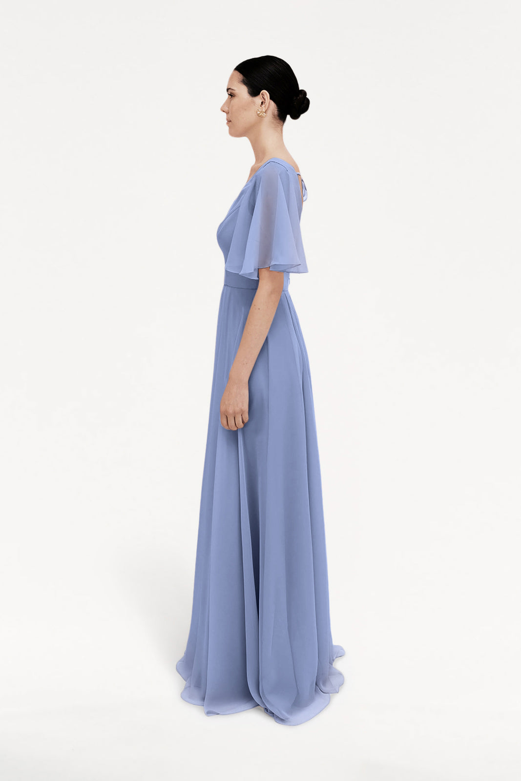 A woman stands in profile, wearing the TH&TH Phoebe Bridesmaid Dress in French Blue chiffon with short flutter sleeves. Her dark hair is styled in a low bun against a plain white background. Ideal as a bridesmaid dress.