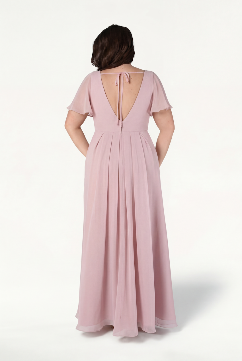 A woman stands against a white background wearing the TH&TH Phoebe Bridesmaid Dress in Smoked Blush Chiffon, featuring short flutter sleeves, a deep V-back, and pockets—a chic, bump-friendly choice.