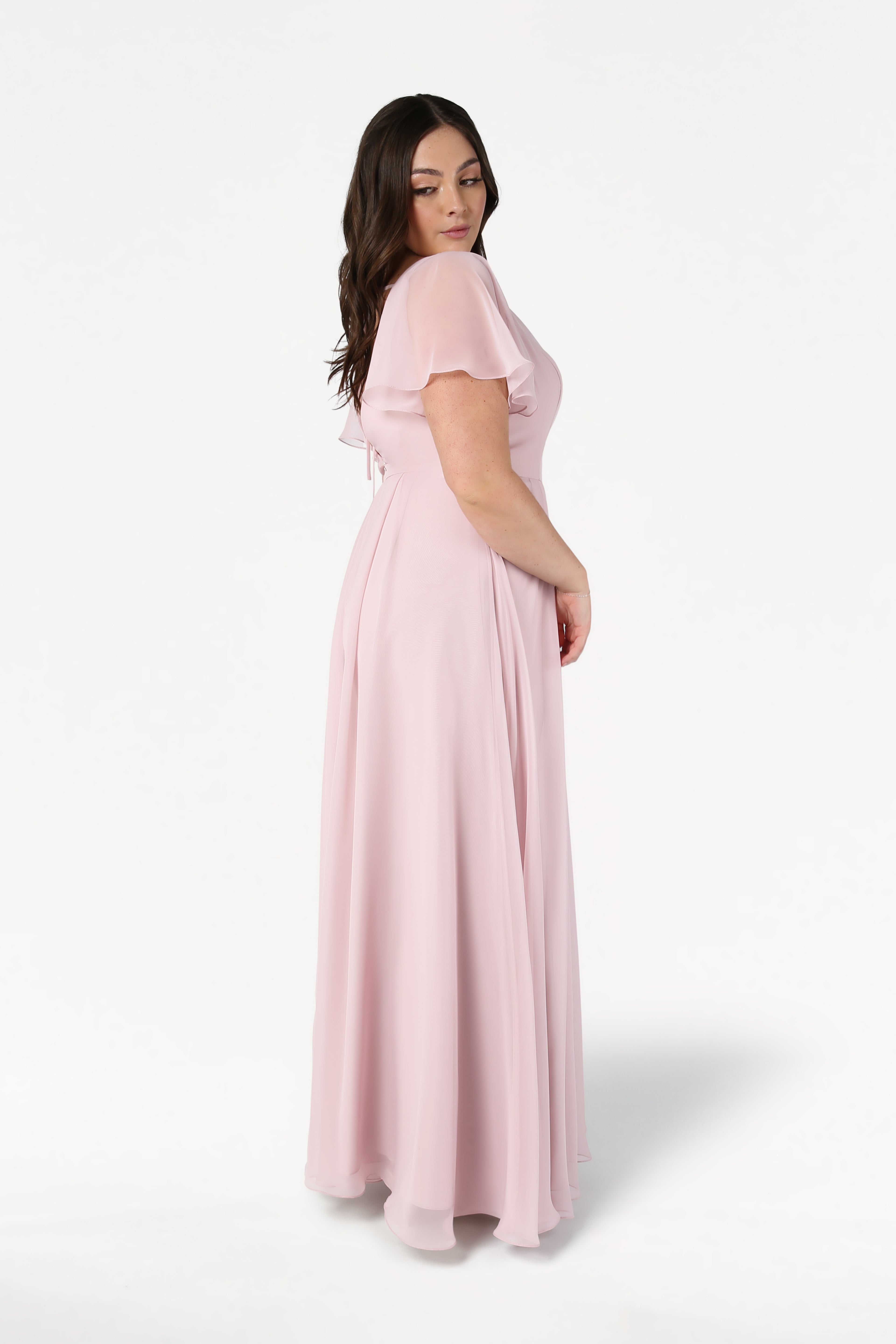 A woman with long dark hair stands sideways in the TH&TH Phoebe Bridesmaid Dress, a floor-length smoked blush chiffon gown with short sleeves, against a plain white background.