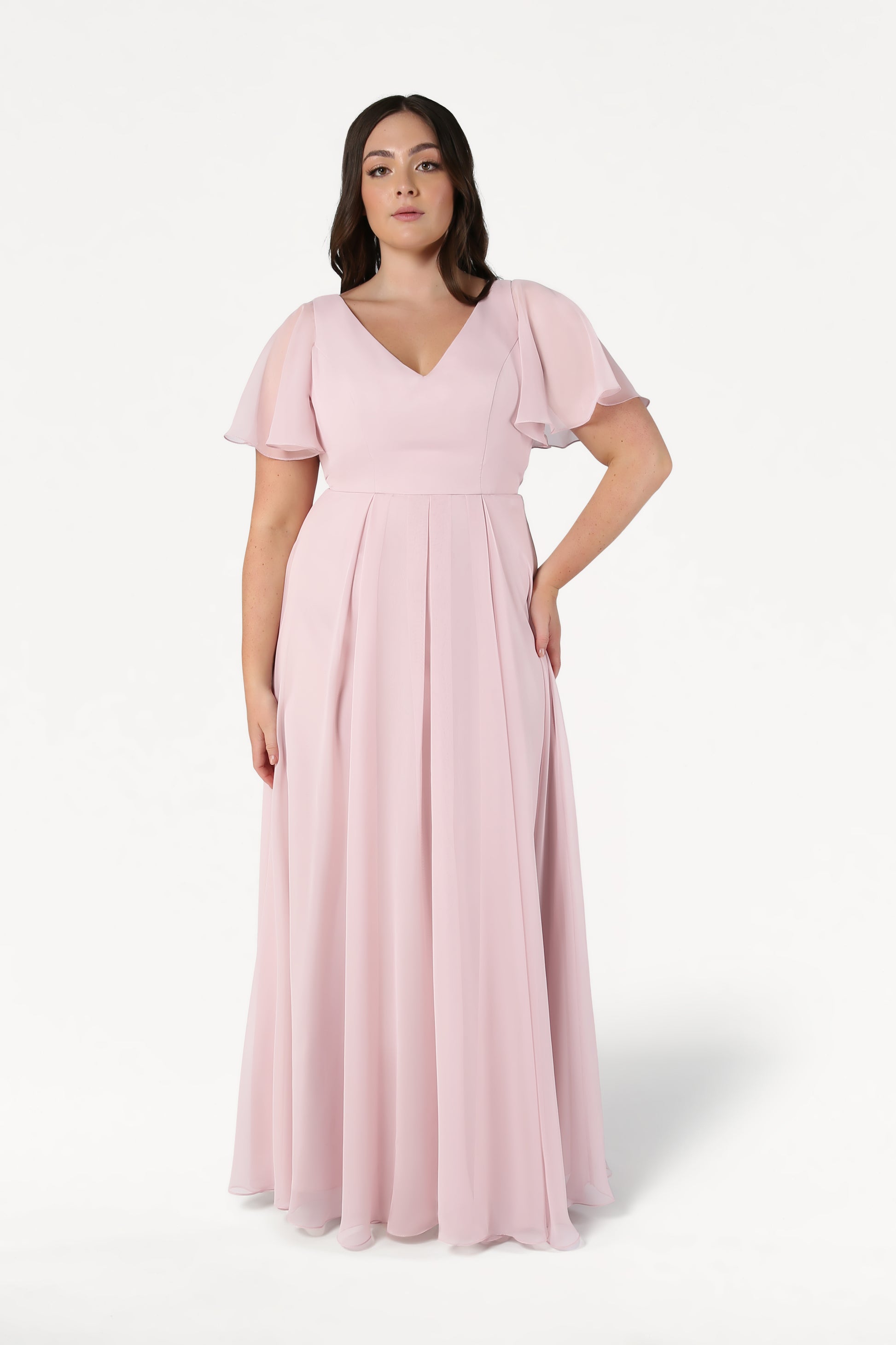 A woman wears the PHOEBE Bridesmaid Dress in Smoked Blush Chiffon by TH&TH, featuring short flutter sleeves and a V-neckline, standing against a white background with a neutral expression—ideal as a bridesmaid dress.