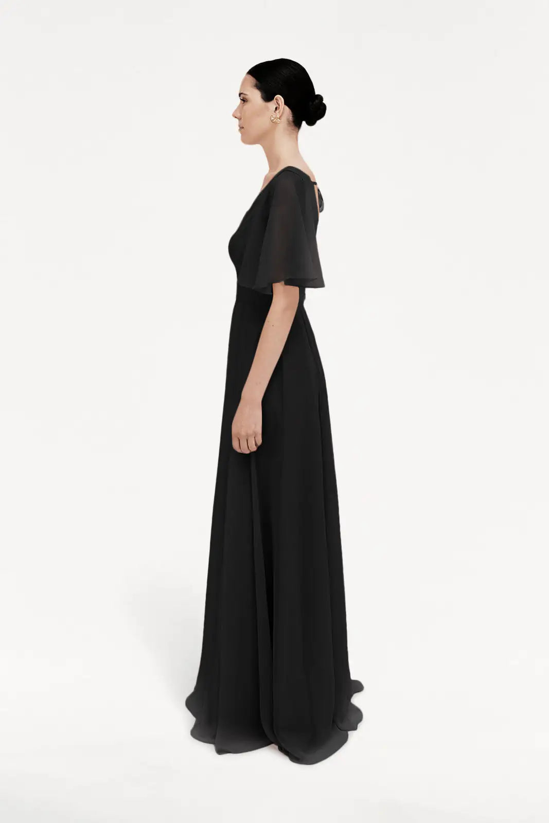 A woman with dark hair in a low bun stands in profile, wearing the TH&TH Phoebe Bridesmaid Dress in Black Chiffon—a floor-length, bump-friendly gown with short sheer sleeves—set against a plain white background.