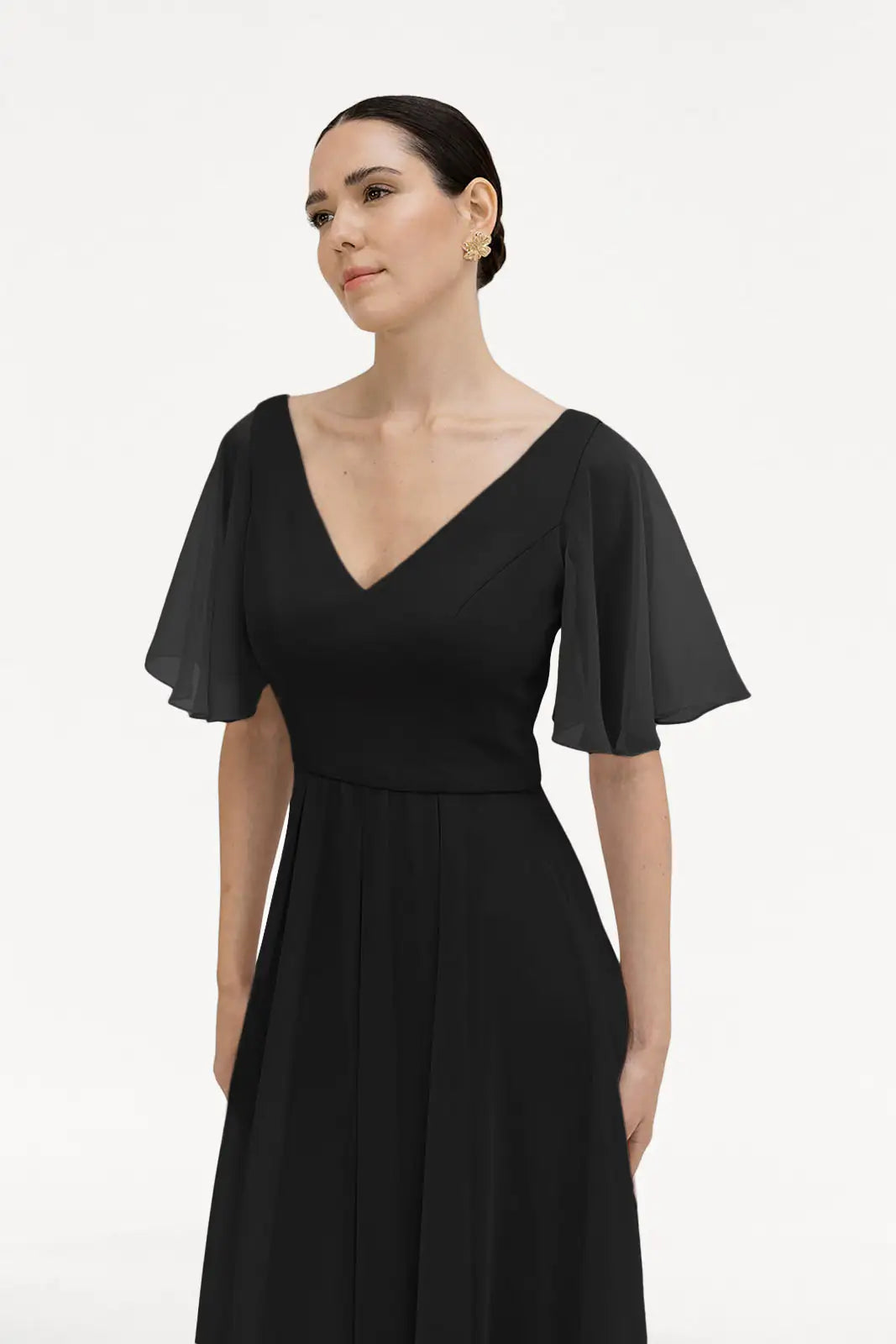 A woman wears the Phoebe Bridesmaid Dress in Black Chiffon by TH&TH—featuring a bump-friendly V-neck, sheer short flutter sleeves, and gold hoop earrings—standing against a plain white background.