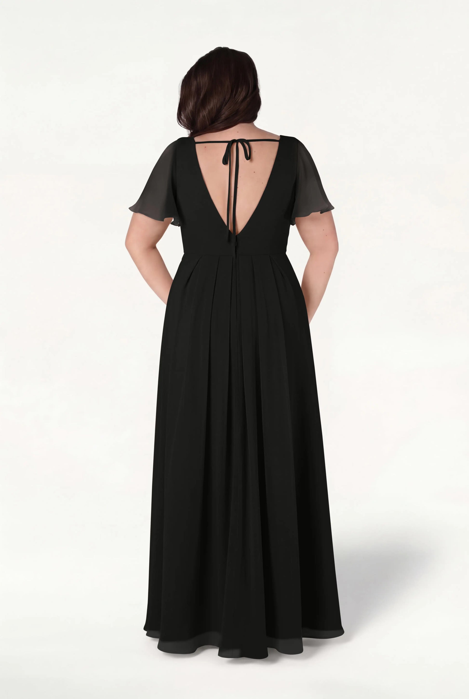 A woman stands facing away on a light background wearing the Phoebe Bridesmaid Dress in Black Chiffon by TH&TH, featuring a floor-length silhouette, short sheer sleeves, and a deep V-back tied at the neck with a ribbon.
