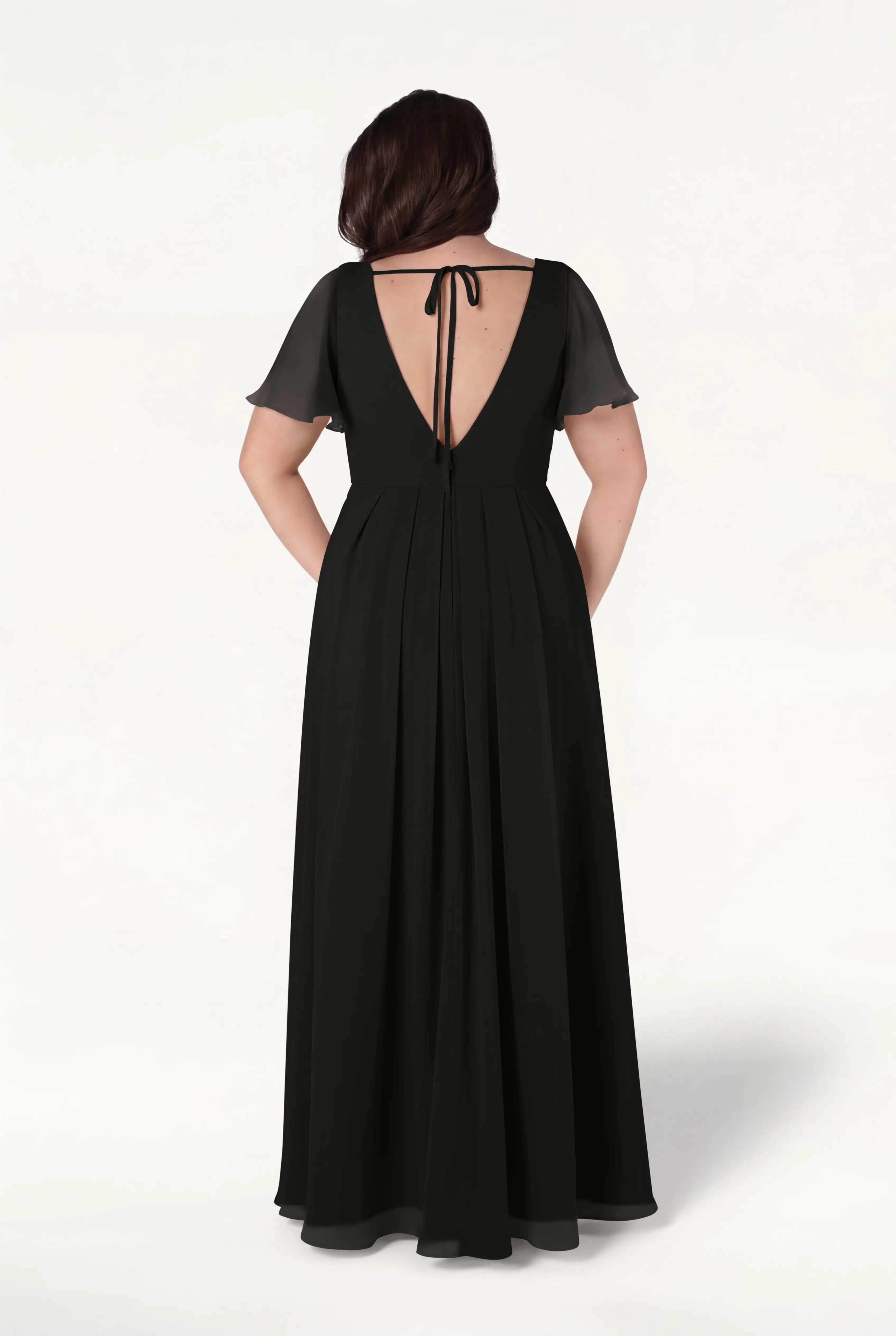 A woman stands facing away on a light background wearing the Phoebe Bridesmaid Dress in Black Chiffon by TH&TH, featuring a floor-length silhouette, short sheer sleeves, and a deep V-back tied at the neck with a ribbon.