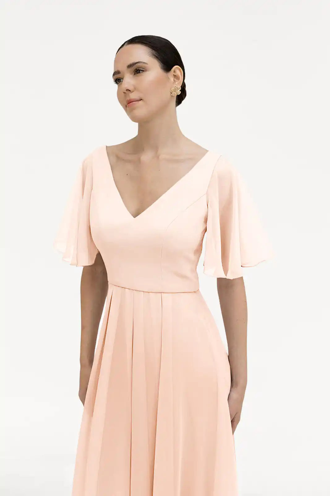 A woman in the TH&TH Phoebe Bridesmaid Dress in blush pink chiffon, featuring a V-neck and flutter sleeves, stands against a white background, looking calmly to the side.