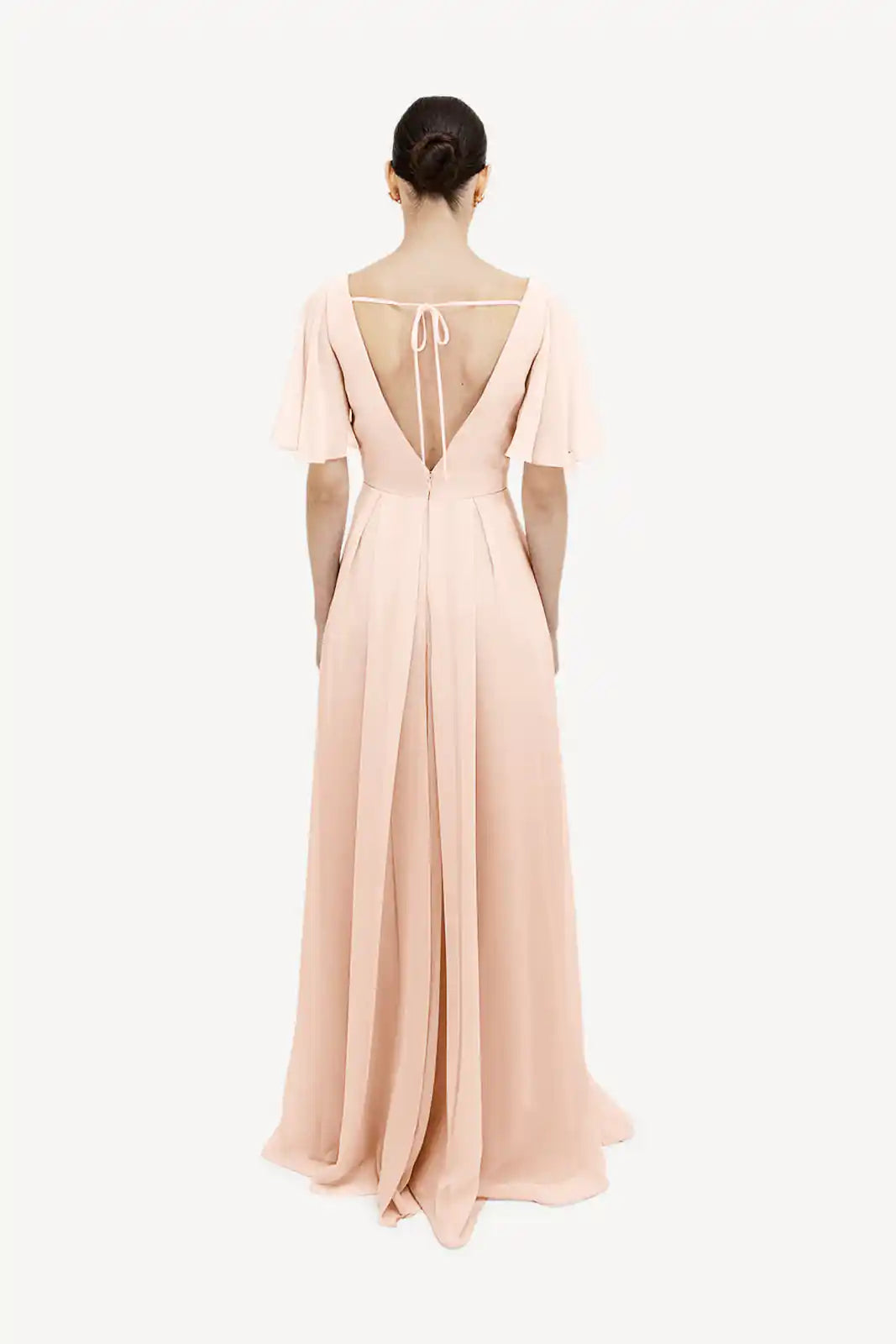 A woman with dark hair in a low bun models the TH&TH Phoebe Bridesmaid Dress in blush pink chiffon, featuring short flutter sleeves and a deep V-back with thin ties, standing against a plain white background.