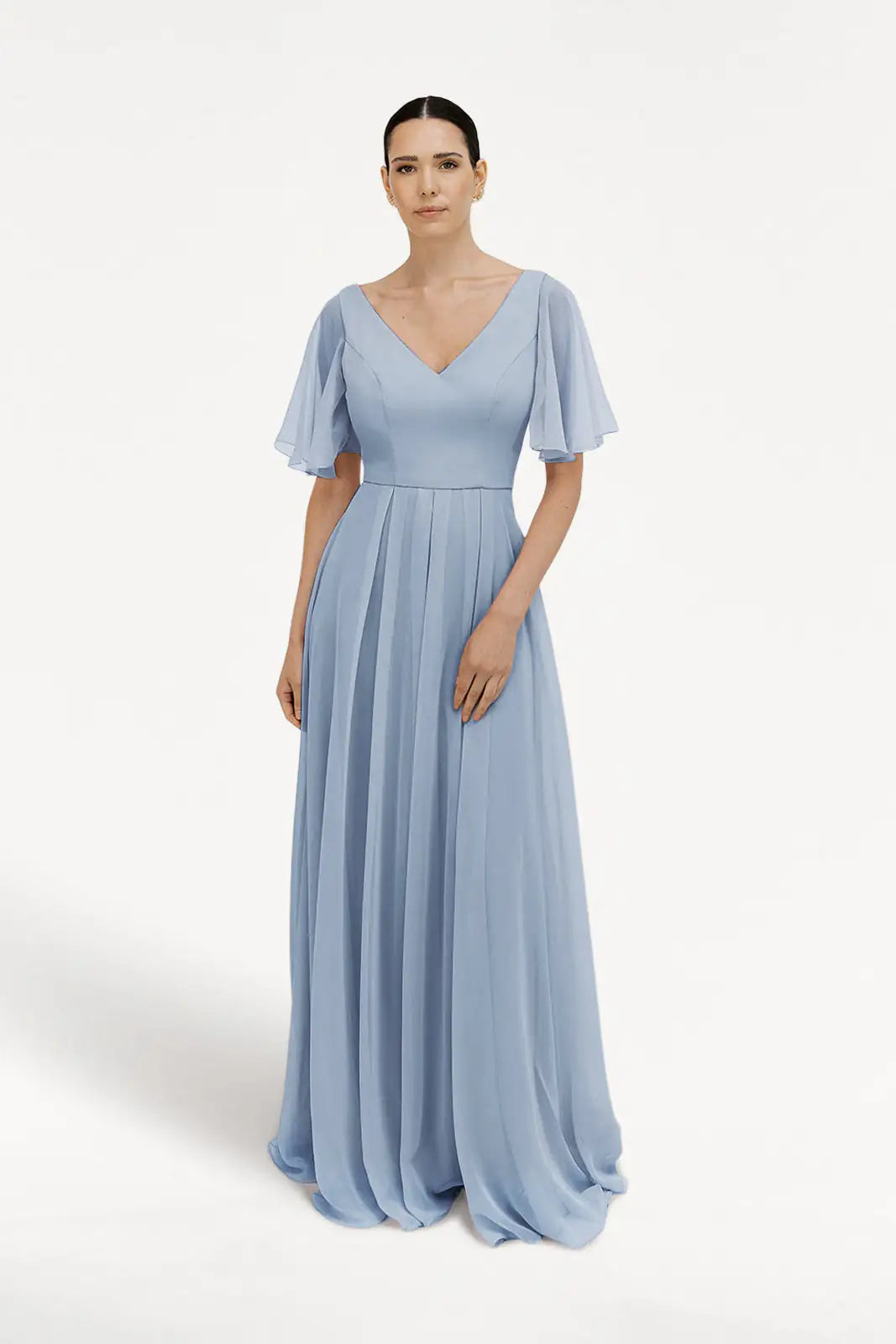 A woman stands against a white background wearing the TH&TH Phoebe Bridesmaid Dress in Dusty Blue Chiffon, featuring short flutter sleeves, a V-neckline, bump-friendly fitted waist, and a floor-length pleated skirt. Her dark hair is pulled back.