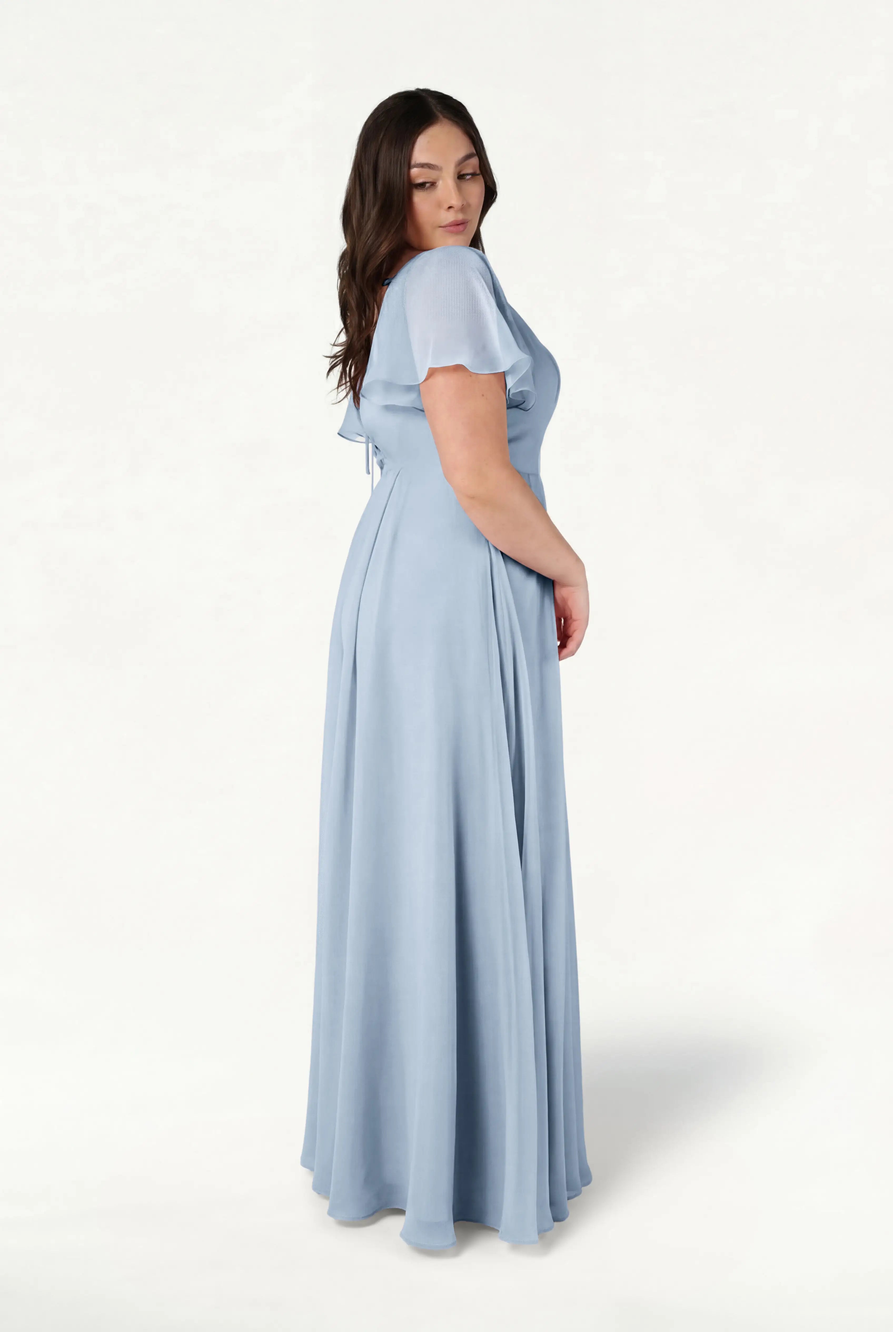 A woman with long brown hair stands sideways, modeling the TH&TH Phoebe Bridesmaid Dress in dusty blue chiffon—featuring a fitted bodice and flowing skirt—against a white background; perfect as a bump-friendly option.