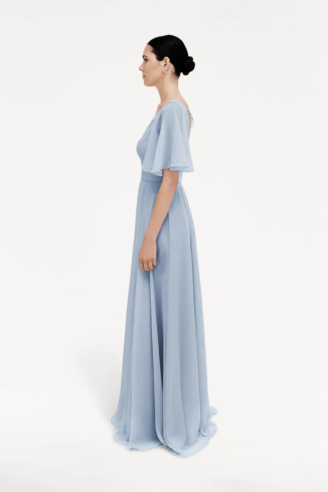 A woman stands in profile against a white background, wearing the TH&TH Phoebe Bridesmaid Dress in dusty blue chiffon with flutter sleeves and her dark hair styled in a neat bun.