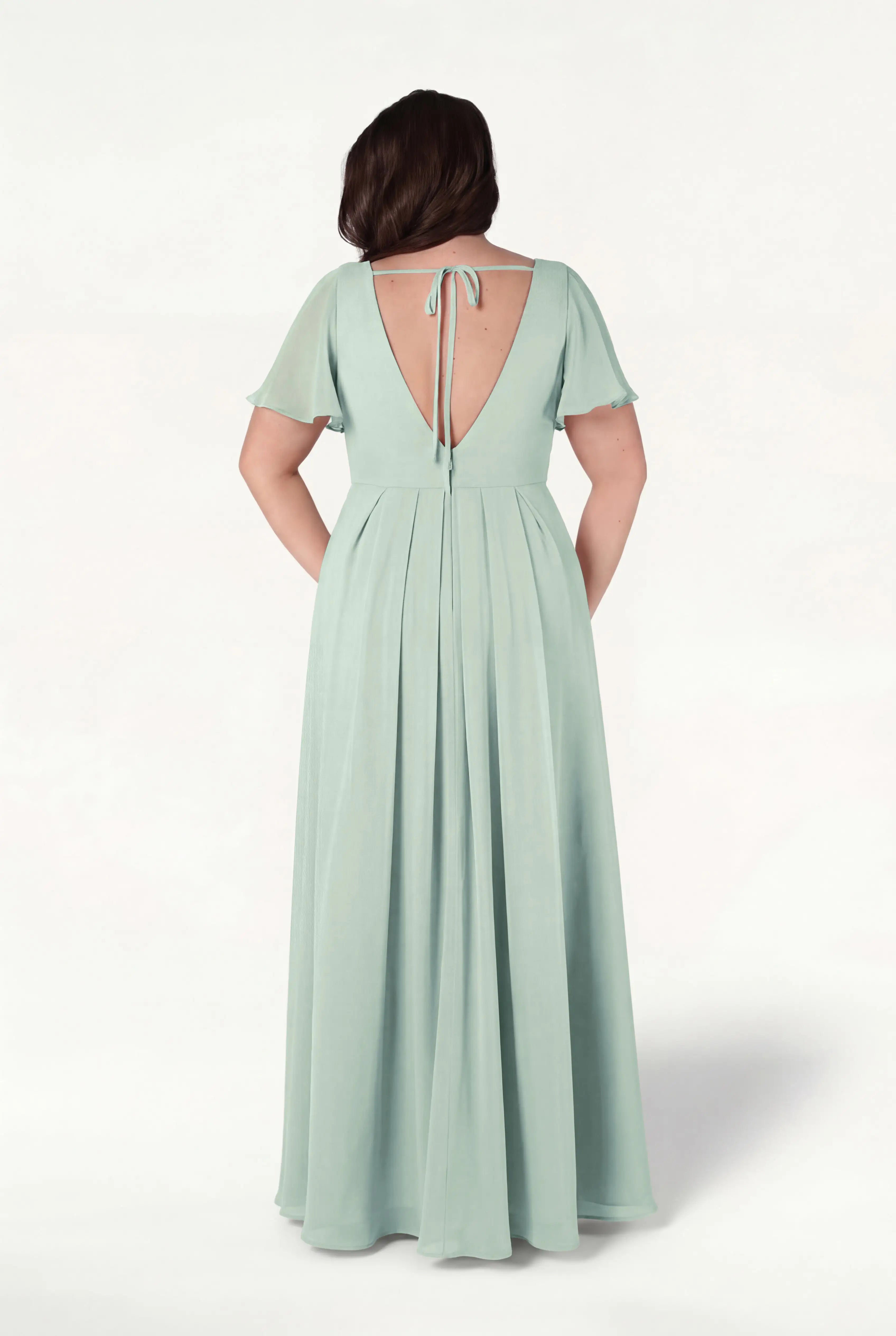 A woman faces away, wearing the TH&TH Phoebe Bridesmaid Dress in Dusty Eucalyptus chiffon—long, flowing with short flutter sleeves, a deep V-back, and tie detail. The background is plain white.