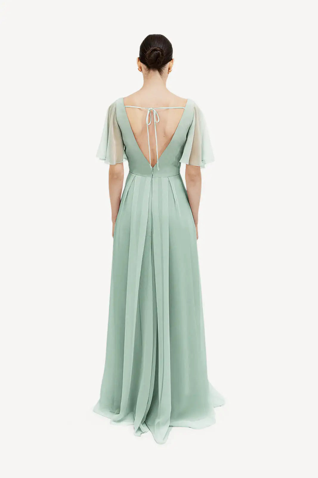 A woman, hair in a neat bun, faces away while wearing the TH&TH Phoebe Bridesmaid Dress in Dusty Eucalyptus chiffon—a floor-length gown with short flutter sleeves, deep V-back, and upper back tie—set against a white background.