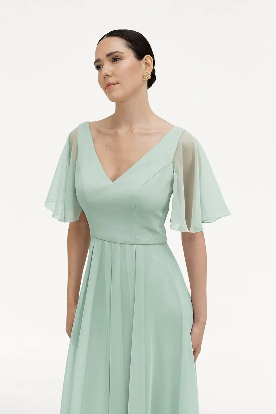 A woman wearing the TH&TH Phoebe Bridesmaid Dress in Dusty Eucalyptus chiffon, featuring sheer flutter sleeves and a V-neckline, stands against a plain white background, looking slightly to the side.