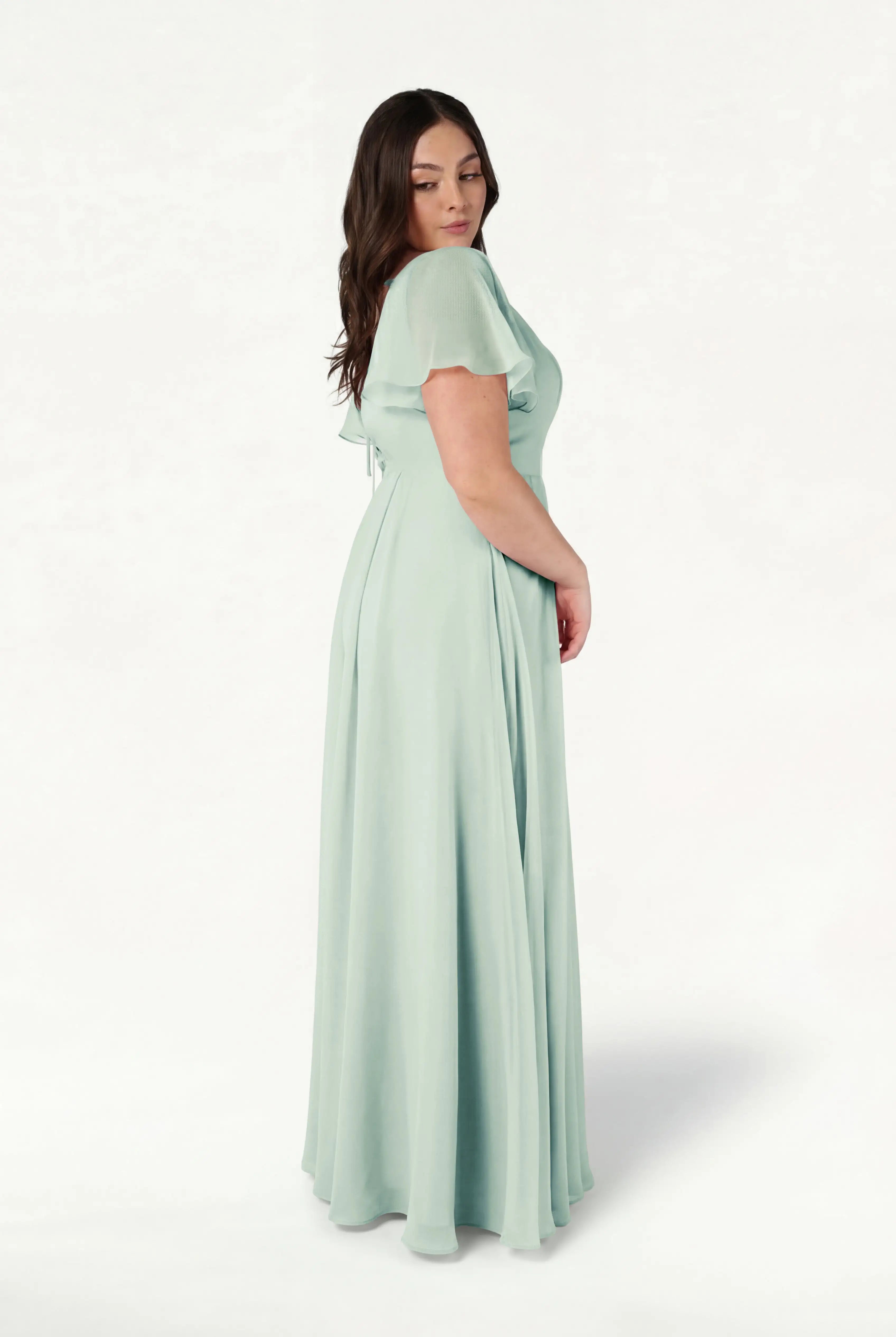 A woman with long dark hair stands sideways, wearing the TH&TH Phoebe Bridesmaid Dress in Dusty Eucalyptus chiffon—a floor-length, bump-friendly gown with a fitted waist and short sleeves—against a plain white background.