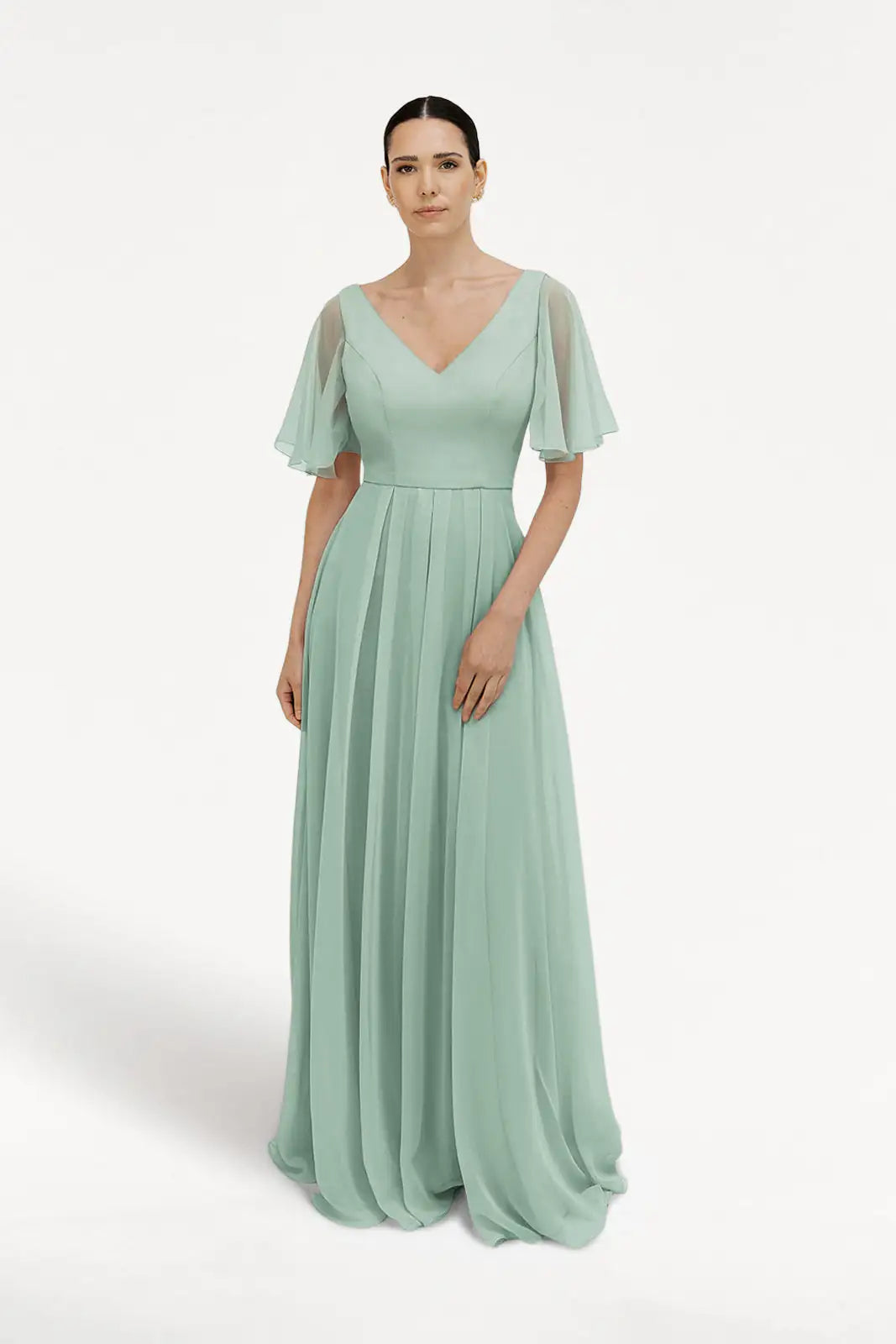 A woman with dark hair pulled back models the TH&TH Phoebe Bridesmaid Dress in Dusty Eucalyptus chiffon, a floor-length gown featuring a V-neckline, sheer flutter sleeves, and a pleated skirt against a white background.
