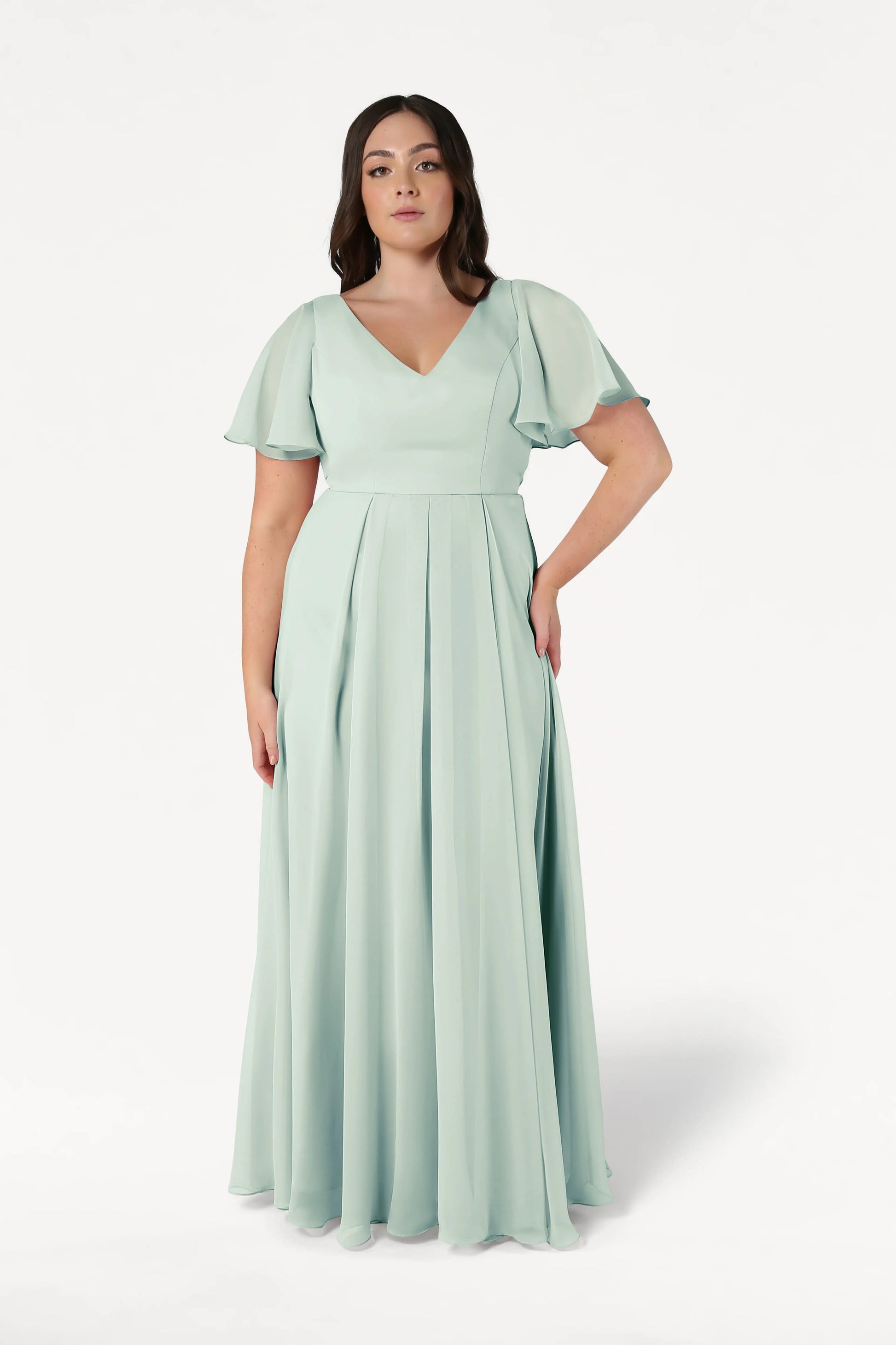 A plus-size model wears the TH&TH Phoebe Bridesmaid Dress in Dusty Eucalyptus chiffon, featuring a floor-length A-line skirt, V-neckline, flutter sleeves, and a fitted bodice.