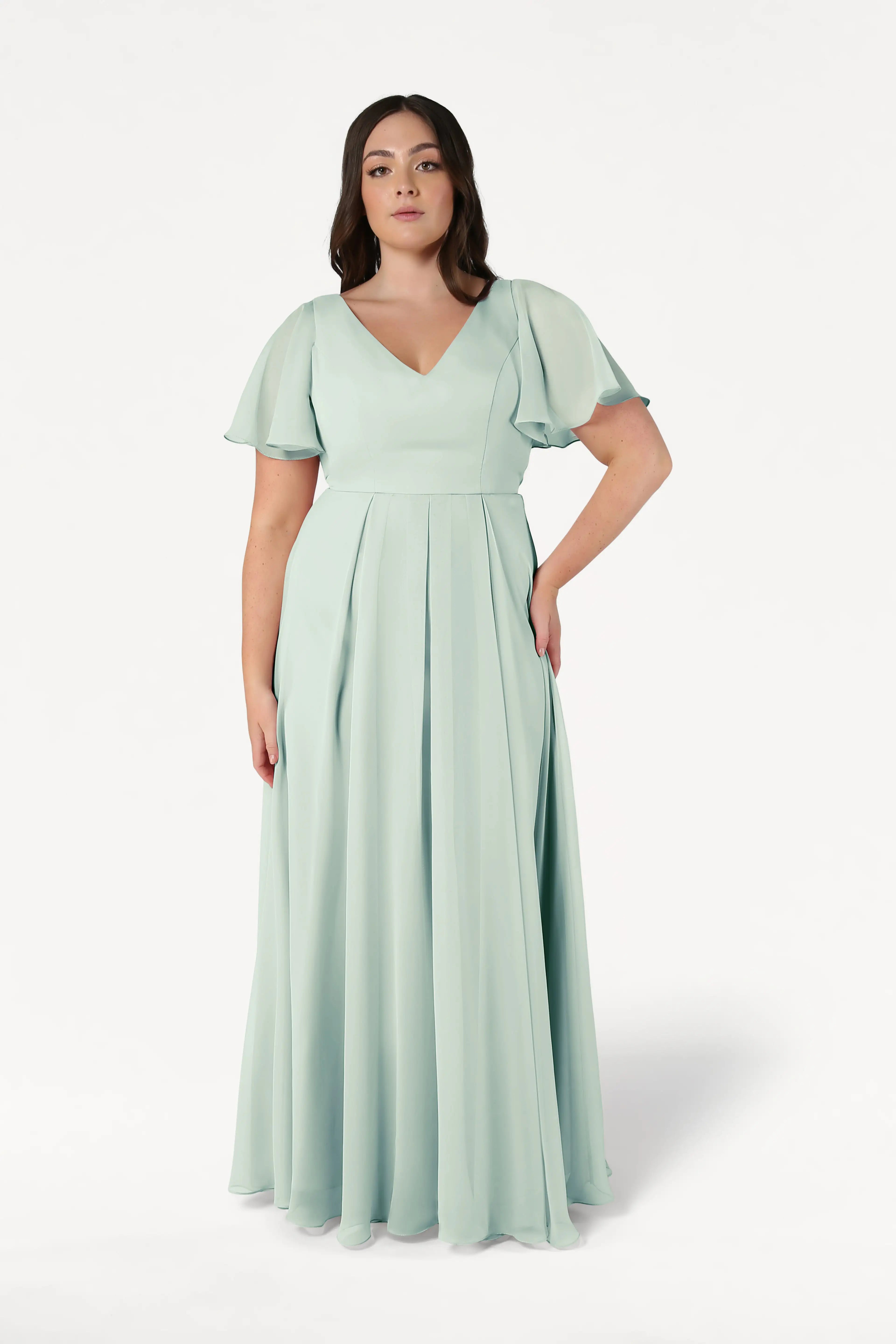 A plus-size model wears the TH&TH Phoebe Bridesmaid Dress in Dusty Eucalyptus chiffon, featuring a floor-length A-line skirt, V-neckline, flutter sleeves, and a fitted bodice.