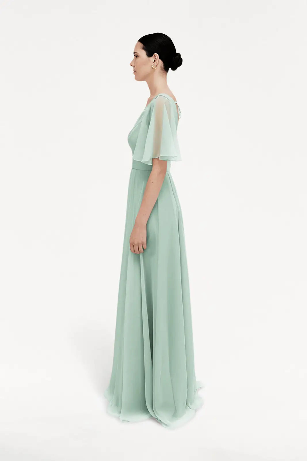A woman stands in profile against a white background wearing the TH&TH Phoebe Bridesmaid Dress in Dusty Eucalyptus Chiffon, featuring short sleeves, a fitted waist, and a sheer overlay.