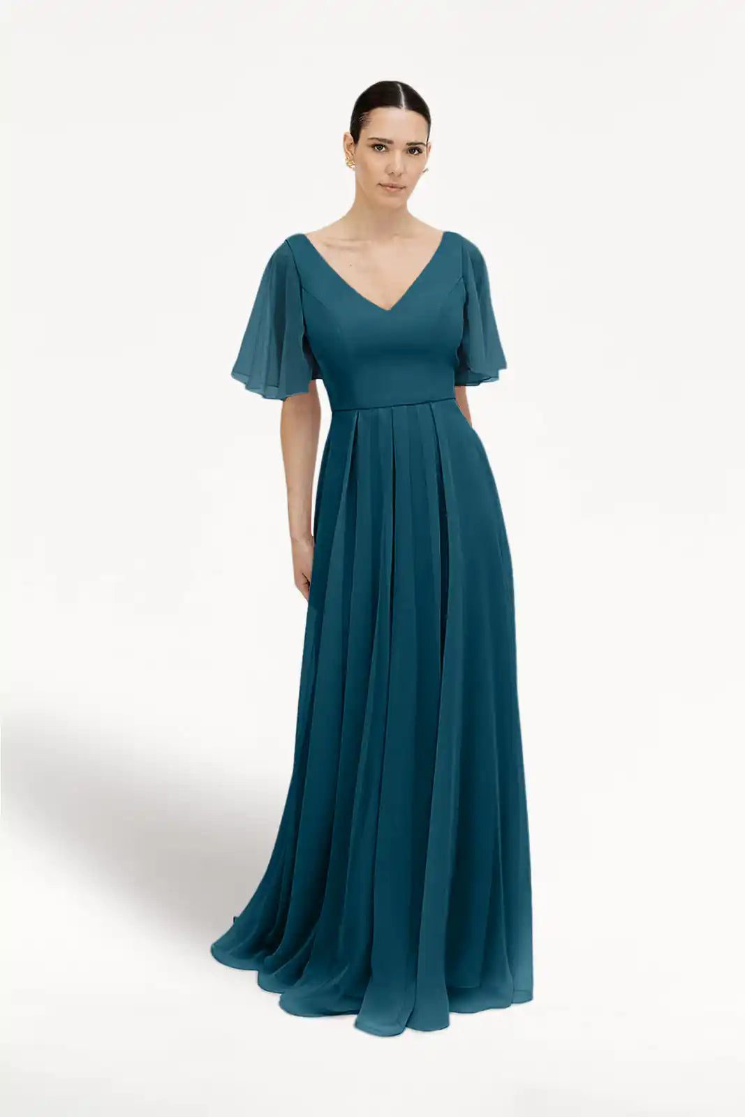 A woman wears the TH&TH Phoebe Bridesmaid Dress in Emerald Dawn Chiffon, a floor-length, bump-friendly gown with flutter sleeves, a V-neckline, fitted bodice, and flowing pleated skirt. The background is plain white.