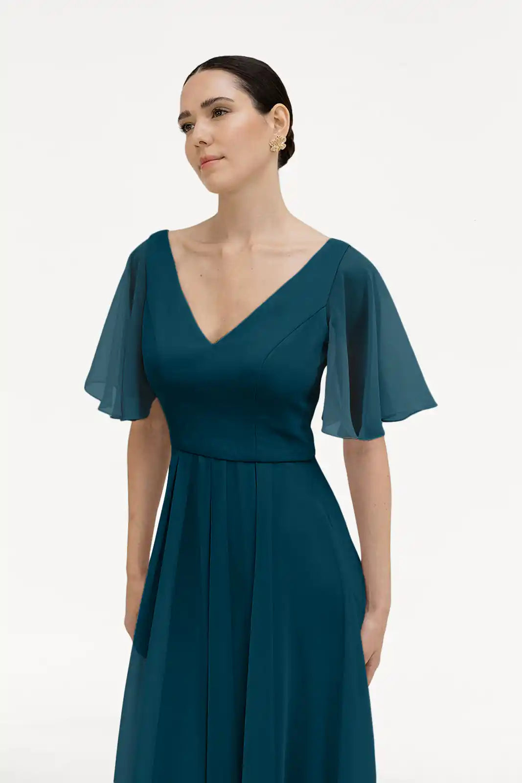 A woman in the Phoebe Bridesmaid Dress in Emerald Dawn Chiffon by TH&TH, featuring a teal V-neck, short sleeves, and sheer flutter sleeves, stands against a white background. This bump-friendly style is ideal for bridesmaids.
