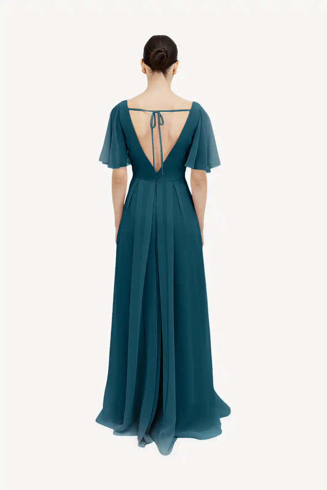 A woman stands with her back turned, wearing the TH&TH Phoebe Bridesmaid Dress in Emerald Dawn chiffon featuring short flutter sleeves, an open back with an upper-back tie, and a neat bun—ideal for a chic bridesmaid look.