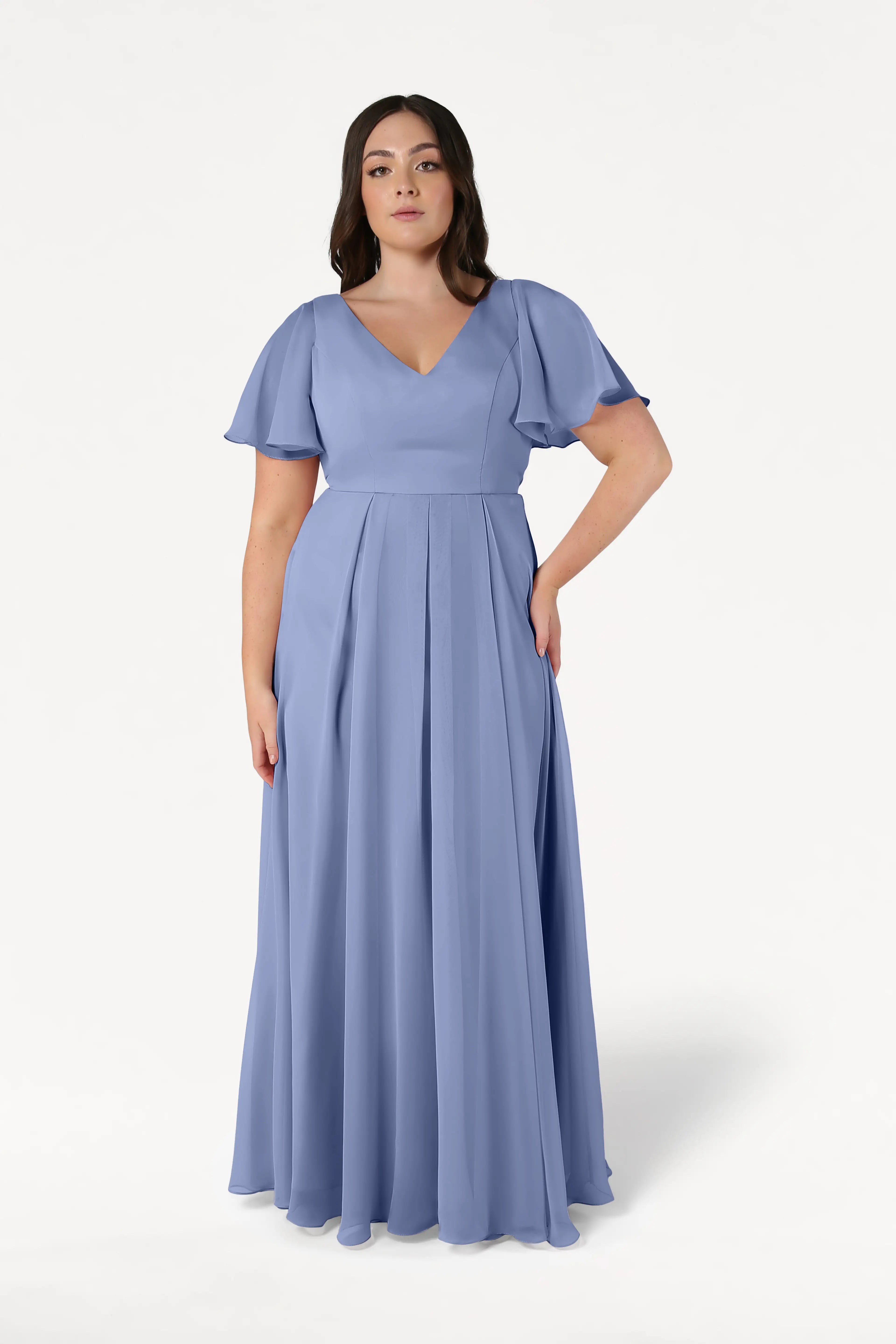 A woman wears the TH&TH Phoebe Bridesmaid Dress in French Blue Chiffon, a floor-length gown with short flutter sleeves and a V-neckline, standing against a plain white background.