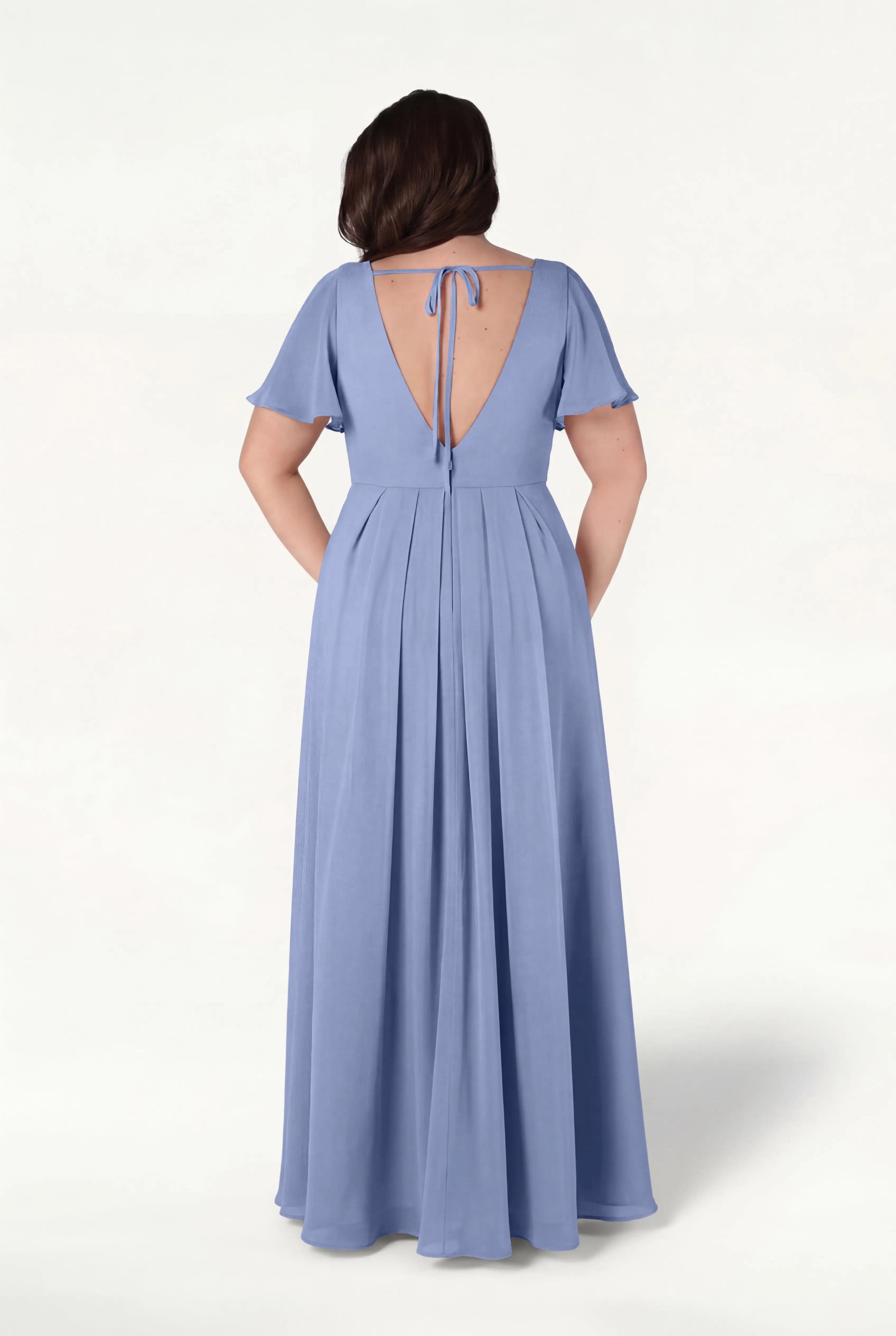 A woman stands against a plain light background, hands in her pockets, wearing the TH&TH Phoebe Bridesmaid Dress in French Blue Chiffon—a floor-length gown with short sleeves and a deep V-back tied at the top with a string.