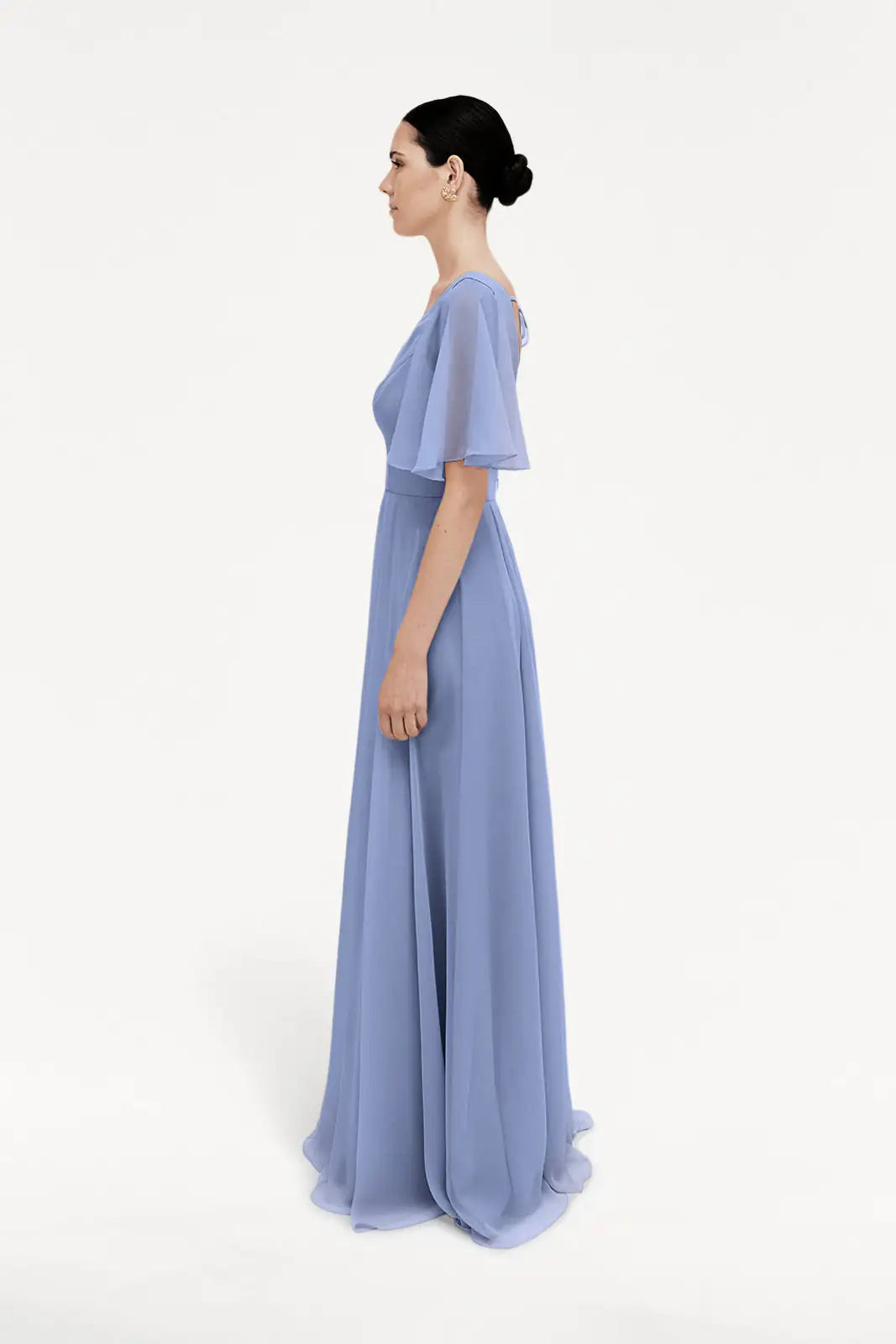 A woman stands in profile, wearing the TH&TH Phoebe Bridesmaid Dress in French Blue chiffon with short flutter sleeves. Her dark hair is styled in a low bun against a plain white background. Ideal as a bridesmaid dress.