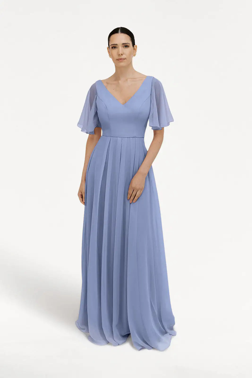 A woman wears the TH&TH Phoebe Bridesmaid Dress in French Blue chiffon, featuring a floor-length pleated skirt, flutter sleeves, and a V-neckline, standing against a plain white background.