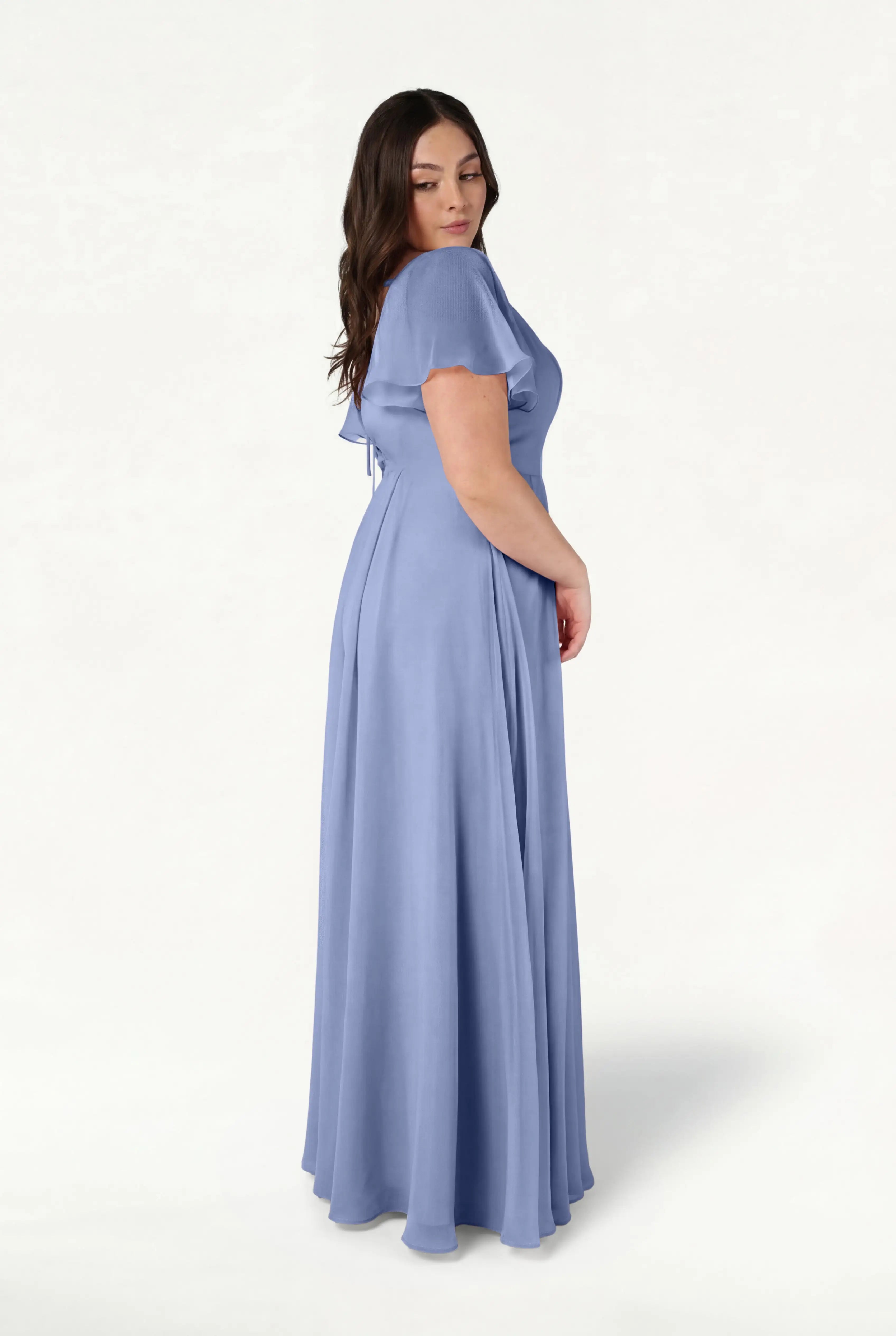 A woman with long brown hair stands sideways against a plain white background, wearing the TH&TH Phoebe Bridesmaid Dress in French Blue Chiffon—a flowing, short-sleeve gown.