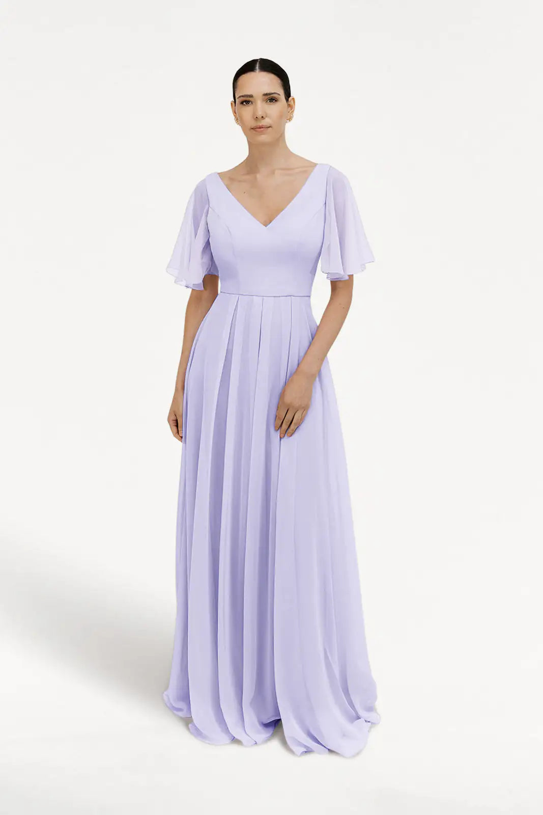 A woman with straight dark hair pulled back wears the TH&TH Phoebe Bridesmaid Dress in Lilac Mist chiffon. The bump-friendly, floor-length gown features flutter sleeves, a V-neckline, fitted waist, and flowing pleated skirt against a white background.